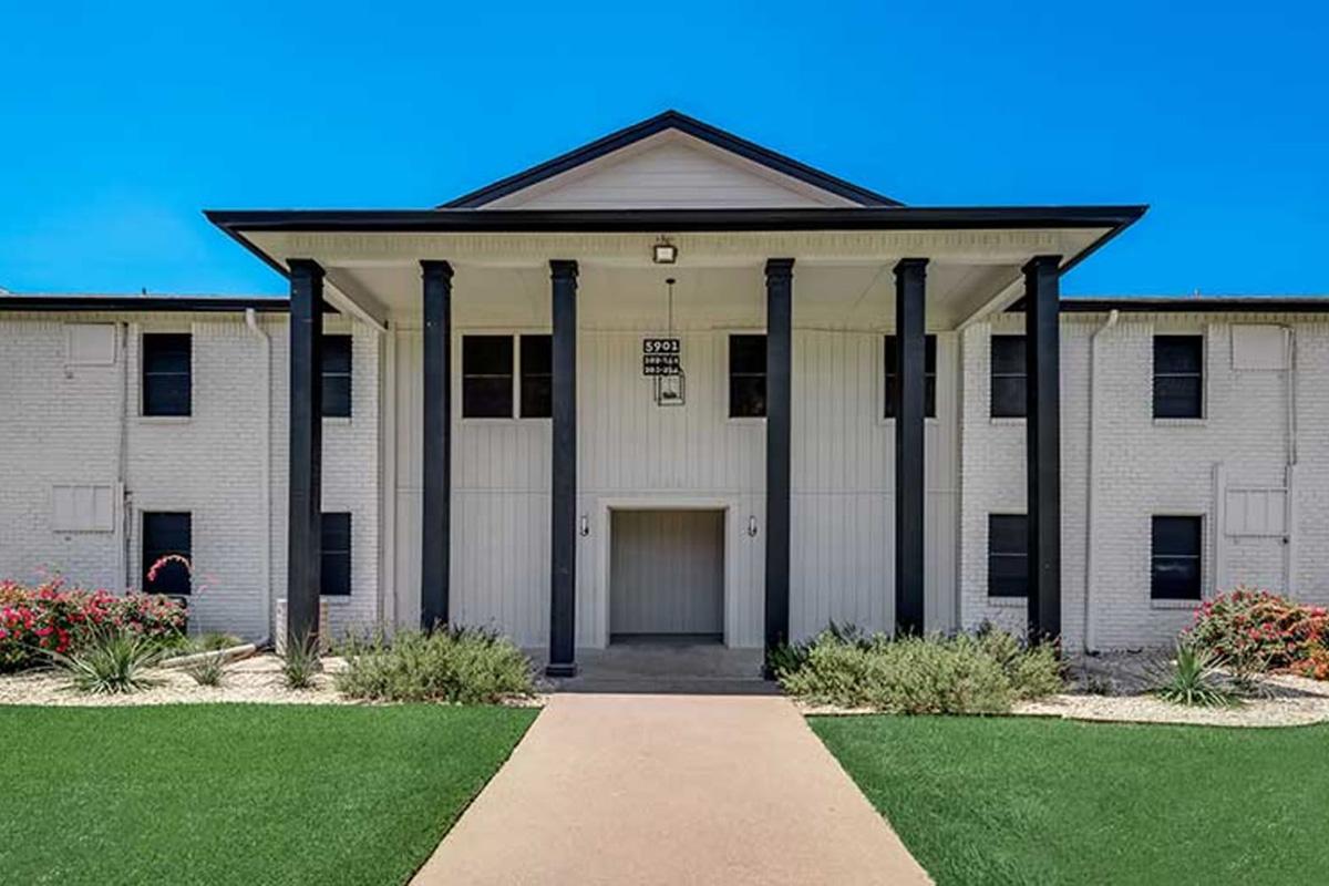 A modern two-story building with a symmetrical facade, featuring four tall black columns at the entrance. The exterior is painted white with decorative shutters. The landscaped front yard includes green grass and ornamental shrubs, and a clear blue sky is visible above.