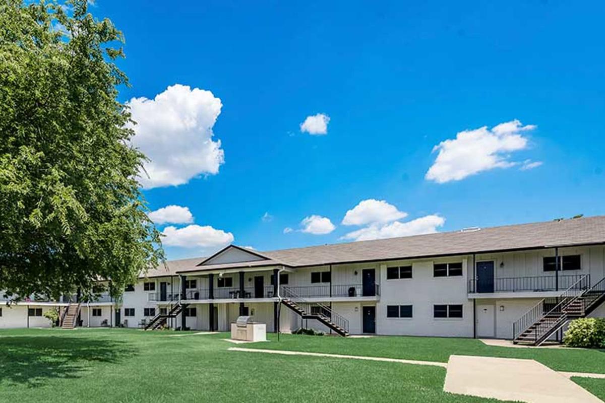 A view of a two-story apartment complex with a grassy lawn and a tree in the foreground. The building features multiple balconies and a walkway. The sky is bright blue with scattered clouds, creating a sunny atmosphere.