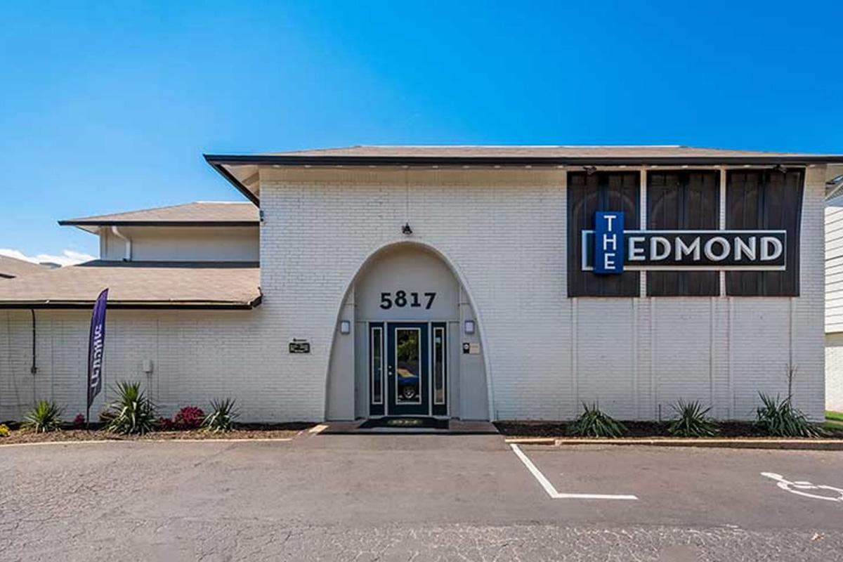 Exterior view of a building labeled "The Edmond." The structure features a light-colored brick facade with a large entrance archway. Signage includes the building name prominently displayed. The surrounding area includes a paved lot with minimal landscaping, and a clear blue sky is visible above.