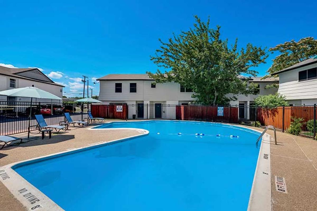 A bright blue swimming pool with lounge chairs and umbrellas arranged around it. In the background, there are residential buildings under a clear blue sky with a few clouds. Lush greenery is visible near the pool area, providing a relaxing atmosphere.