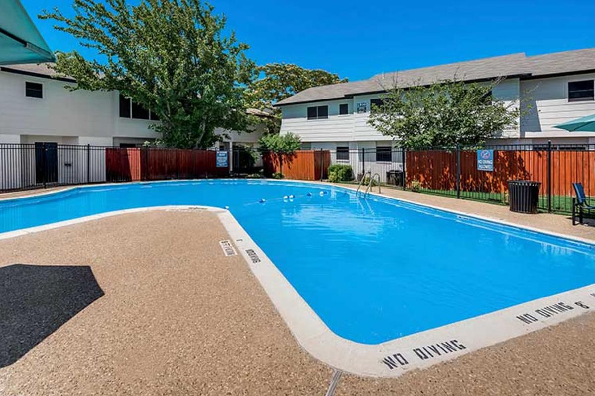 A clear blue swimming pool surrounded by a landscaped area, with nearby buildings visible. The pool deck features a "No Diving" sign, and chairs are placed at the edges. Lush trees and shrubs frame the scene under a bright, sunny sky.