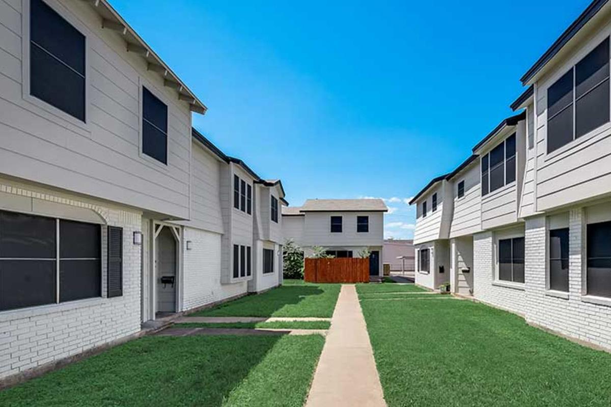 A view of a well-maintained courtyard featuring two rows of white buildings with dark windows. The grassy pathway runs between the buildings, leading towards a single structure at the back. The sky is clear and blue, enhancing the bright, open atmosphere of the neighborhood.