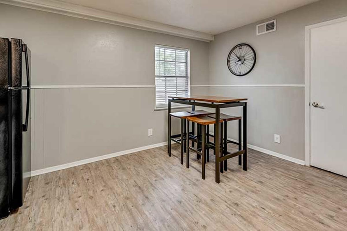 A modern kitchen featuring a black refrigerator, light gray walls, and wooden flooring. There’s a small dining area with a high table and two stools. A wall clock hangs above the table, and a window provides natural light, enhancing the open and airy feel of the space.