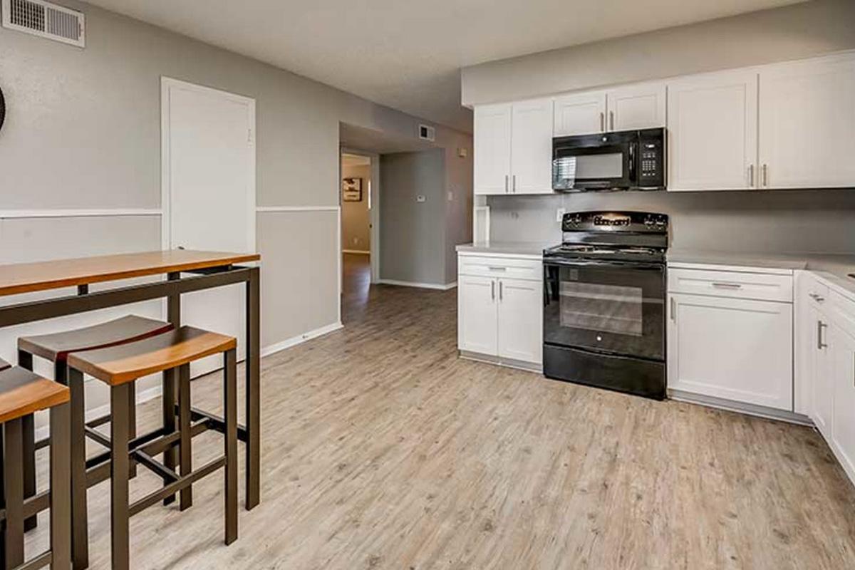 A modern kitchen featuring white cabinetry, a black stove and microwave, and a wooden dining table with bar stools. The floor is a light wood laminate, and the walls are painted in a soft gray. A doorway leads to another room in the background.