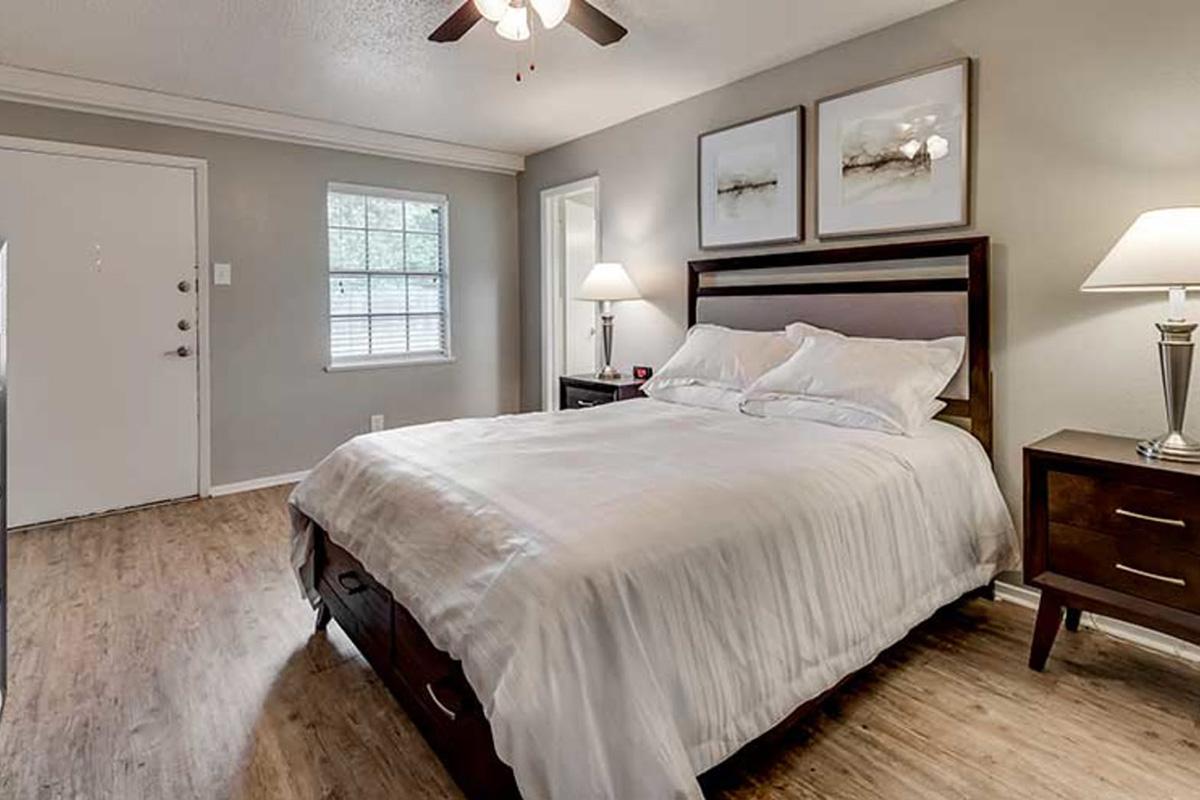 A cozy bedroom featuring a queen-sized bed with white linens, a large window with blinds, and two framed pictures above the bed. There's a bedside table with a lamp on each side of the bed, and wooden flooring complements the neutral-colored walls. A doorway leads to another room, and a front door is visible at the back.