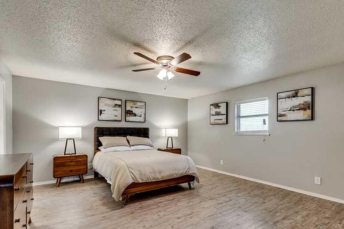 A spacious, modern bedroom featuring a wooden bed with white bedding, two bedside lamps on matching nightstands, and three abstract paintings on the wall. Natural light comes through a window, and a ceiling fan adds to the room’s comfortable atmosphere. The flooring is light-colored wood.