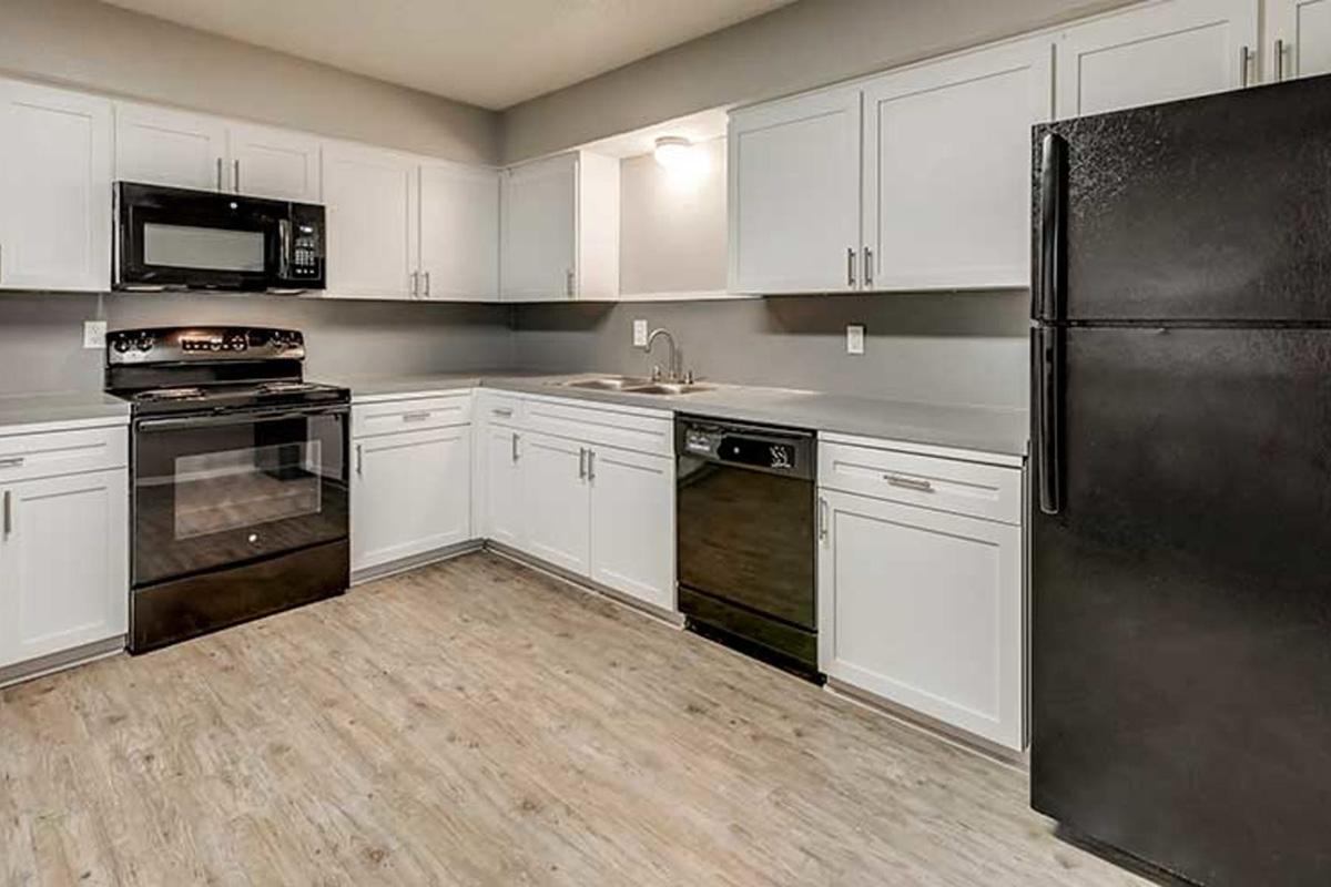 A modern kitchen featuring white cabinets, a black refrigerator, an oven, a microwave, and a dishwasher. The countertops are light gray, and the flooring is a wood-like material. The kitchen is well-lit with overhead lights, creating a bright and inviting atmosphere.