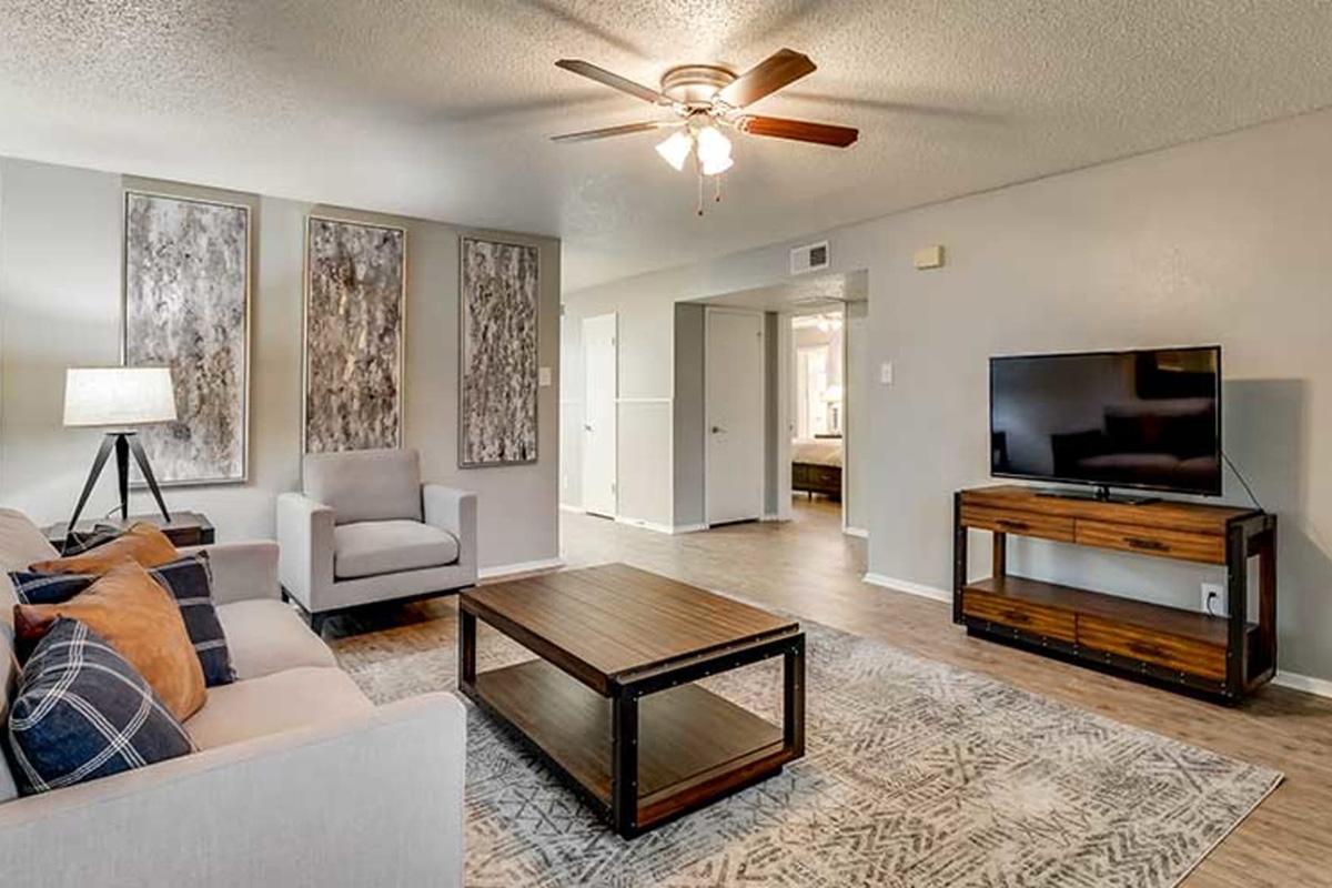 A cozy living room featuring a light-colored sofa with decorative pillows, a wooden coffee table, and a flat-screen TV on a console. The space has textured wall art, a ceiling fan, and soft lighting from a floor lamp. The décor exudes a modern and inviting atmosphere.