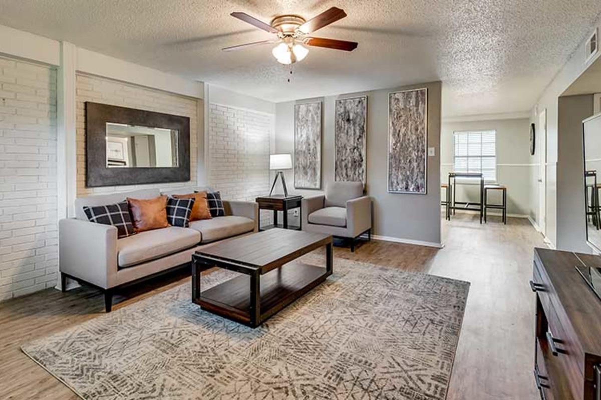 Living room featuring a light gray sofa with orange and black pillows, a wooden coffee table, and a decorative area rug. The walls are painted in neutral tones, with a large mirror above the sofa and two abstract wall art pieces. A lamp stands beside the sofa, and there is a glimpse of a dining area in the background.