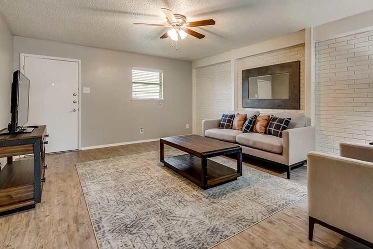 A cozy living room featuring a light-colored sofa with patterned cushions, a wooden coffee table, and an area rug with geometric designs. There is a flat-screen TV on a wooden stand, a ceiling fan, and a window providing natural light, creating a warm and inviting atmosphere.