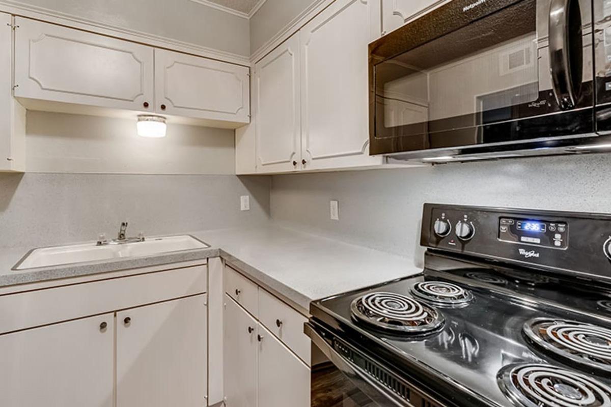 A modern kitchen featuring white cabinetry, a stainless steel microwave above a black stovetop, and a sink with a single light fixture above. The countertop is gray, with a clean and organized appearance, showcasing an efficient use of space.