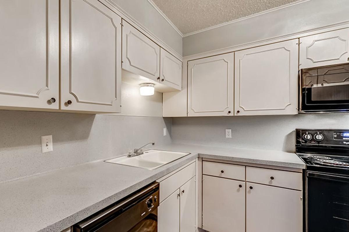 A modern kitchen with white cabinetry and a light gray countertop. It features a sink, a stainless steel oven, and a black microwave. The walls are painted light gray, and the cabinets have decorative paneling, creating a clean and inviting atmosphere.