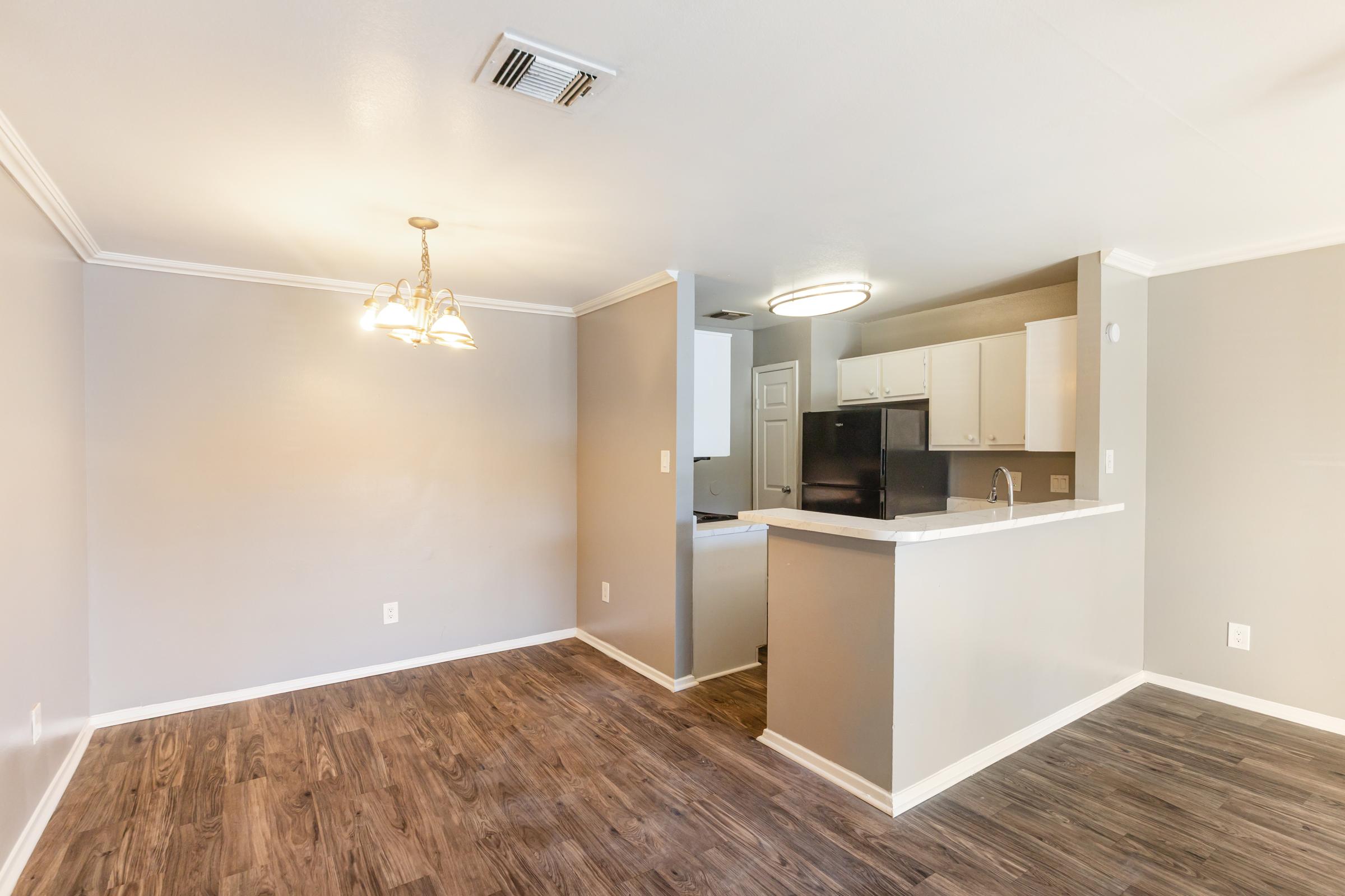 Interior view of a modern apartment featuring a light-filled living area connected to a kitchen. The space has hardwood-style flooring, neutral gray walls, a chandelier above the dining area, and white cabinetry in the kitchen. The layout is open and inviting with natural light from the window.