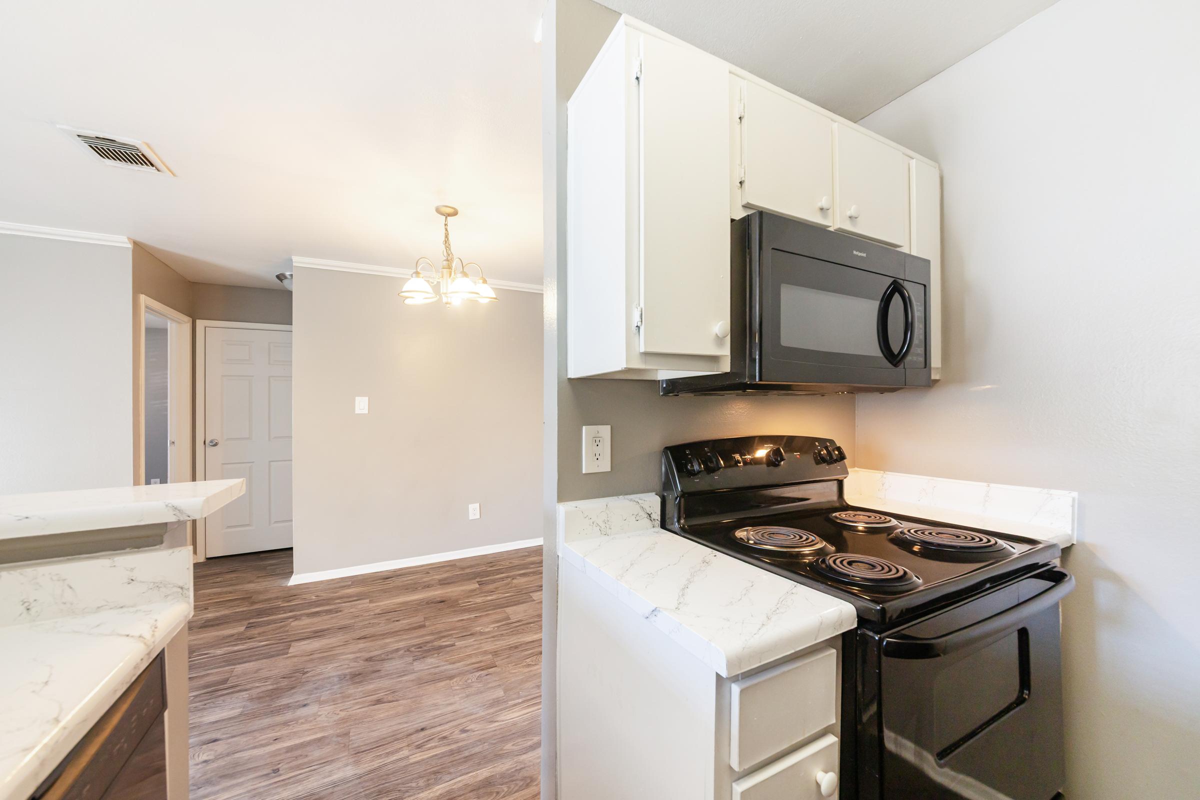 A modern kitchen featuring a black electric stove and microwave, white cabinets, and light countertops. The flooring is a warm-toned wood laminate. In the background, a dining area with a chandelier is visible, along with a doorway leading to another room. Soft, neutral wall colors enhance the inviting atmosphere.