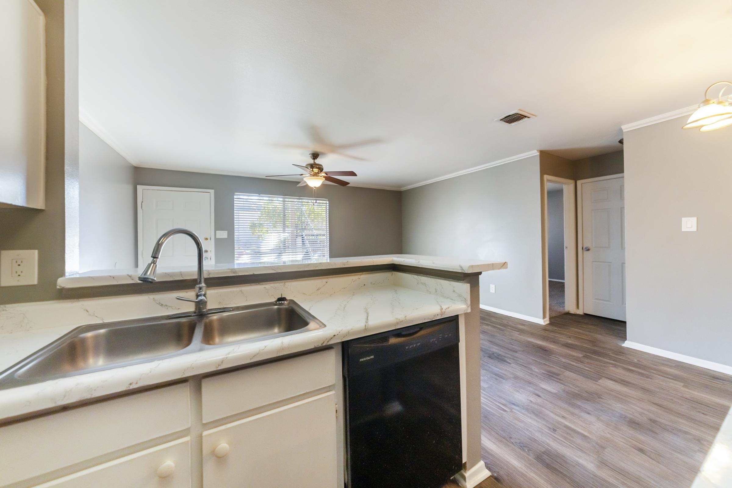 A view of a modern kitchen featuring a white countertop with a double sink, a black dishwasher, and a ceiling fan. The open layout shows a light gray living space with a window and doors leading to other rooms, complemented by warm wood-like flooring.