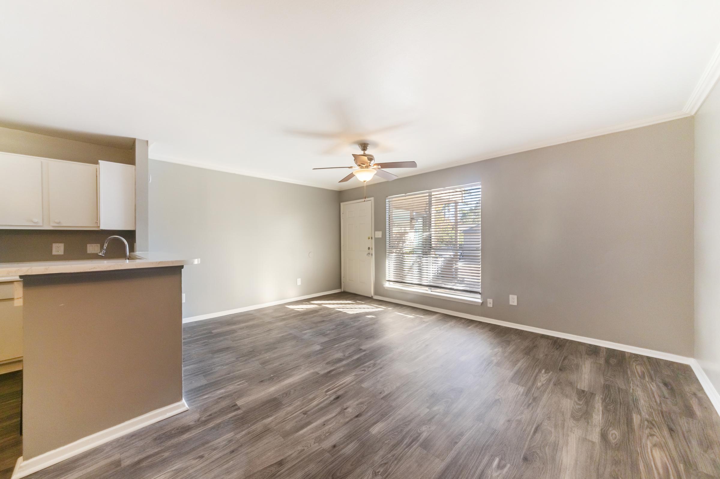 A spacious living area with light-colored walls and wooden flooring. A ceiling fan is overhead, and a window with blinds allows natural light to enter. To the left, there is a simple kitchen area with white cabinets. The room appears bright and open, creating a welcoming atmosphere.