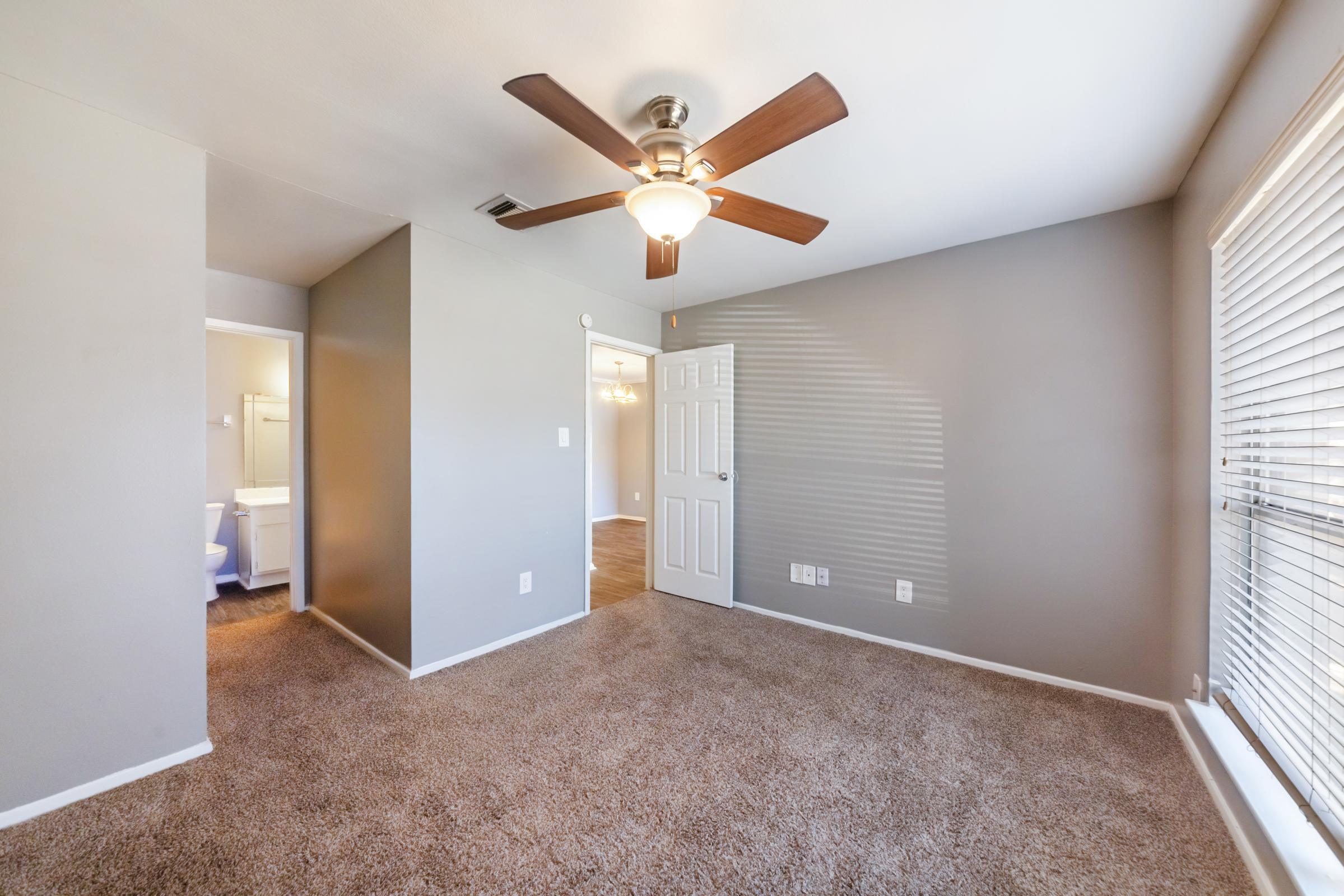 A spacious room with light gray walls and carpet, featuring a ceiling fan with a light fixture. The room has a doorway leading to a bathroom on the left and another room visible at the end. Natural light filters in through the blinds on a nearby window, creating a bright and airy atmosphere.