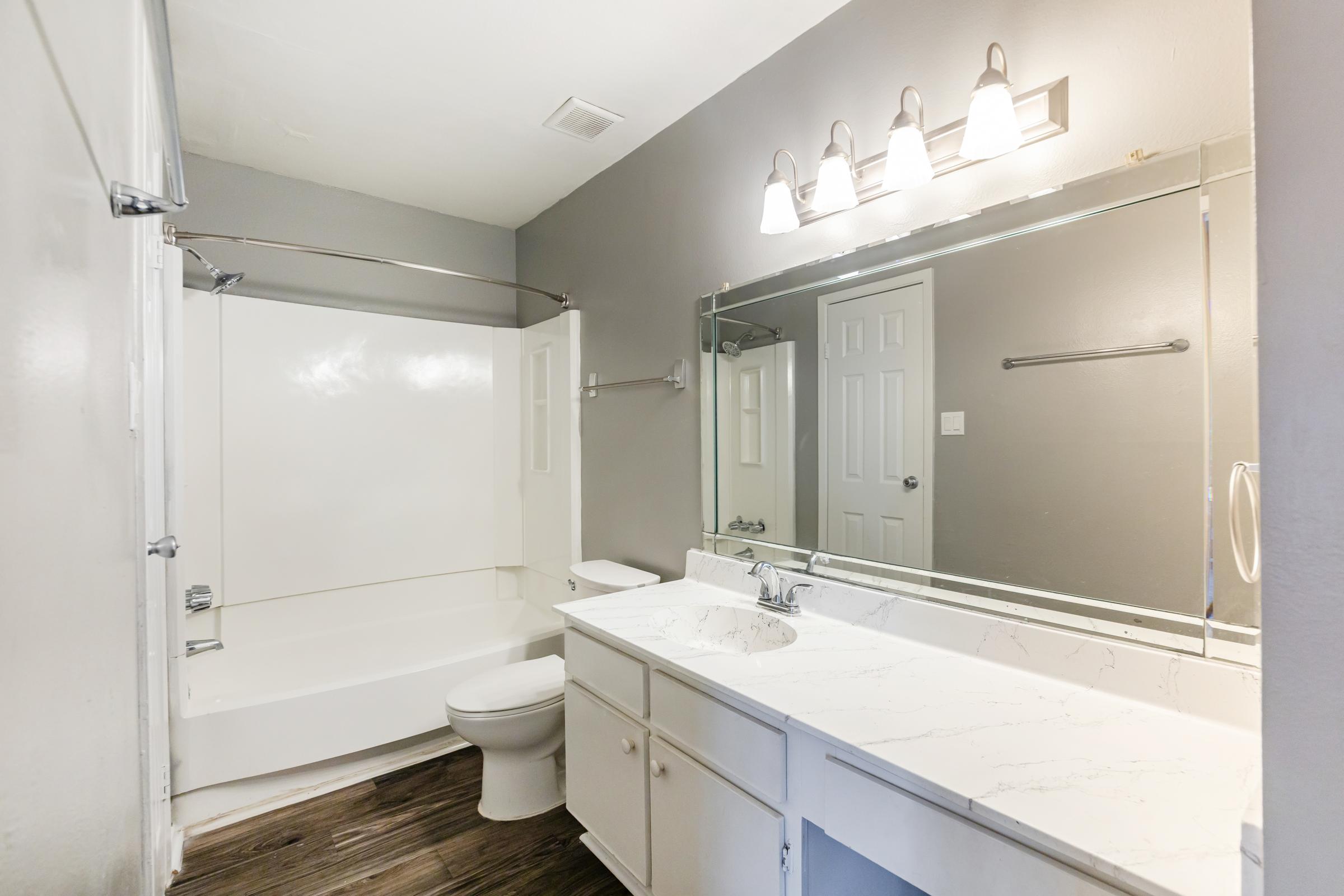 A modern bathroom featuring a white bathtub with a shower curtain, a white toilet, and a double sink vanity with a marble countertop. The walls are painted gray, and there are three light fixtures above the mirror. A door is visible in the background, adding to the spacious feel of the room.