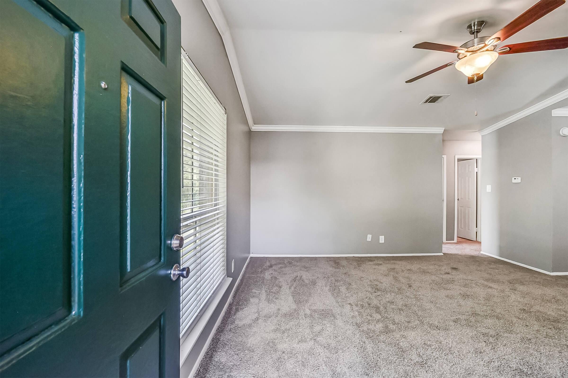 View from an open green front door into a spacious living room. The room features light gray walls, plush carpet, a ceiling fan, and a window with blinds. A doorway on the right leads to another room, enhancing the open and airy feel of the space.