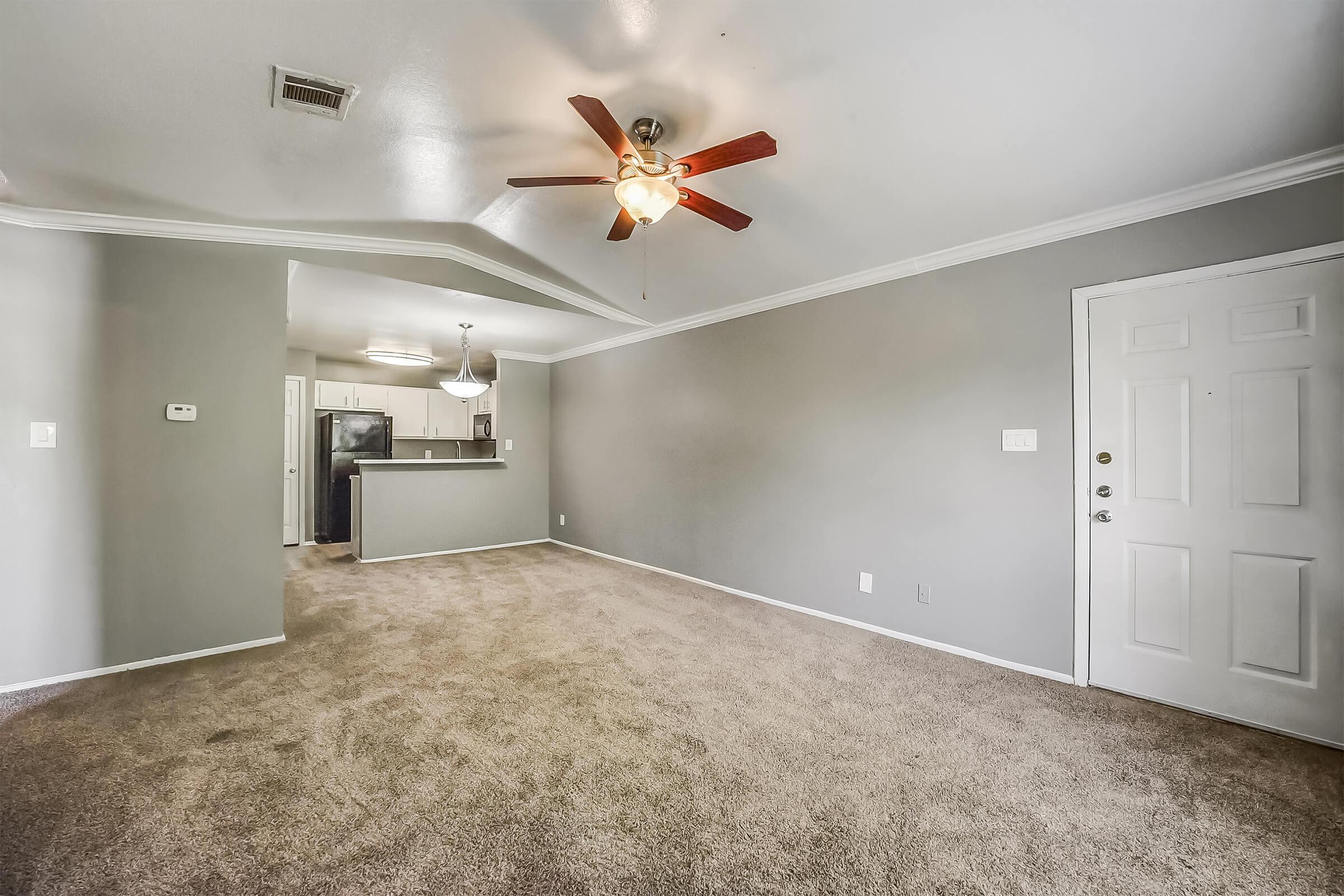 A spacious living room featuring light gray walls, a ceiling fan with wooden blades, and beige carpet. A doorway leads to an entrance, while an open kitchen area is visible in the background with modern appliances. Soft lighting fixtures illuminate the space, creating a warm atmosphere.
