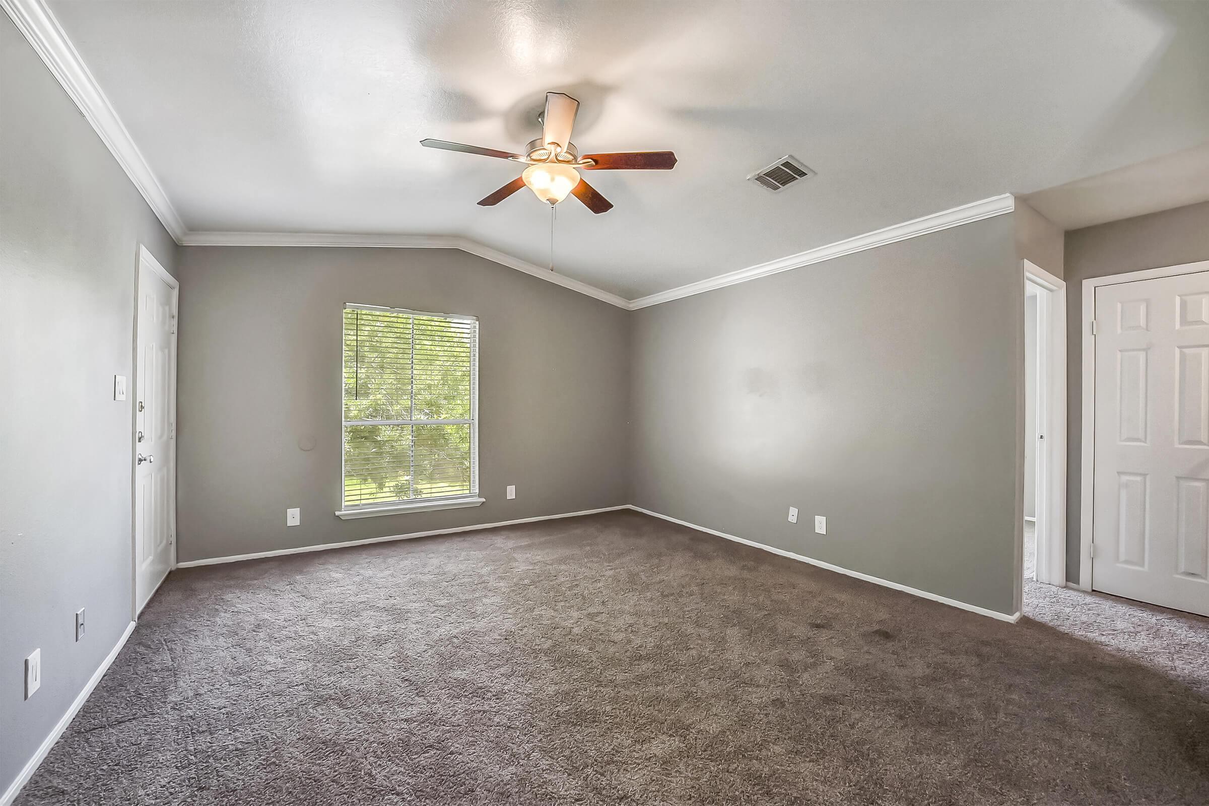 A spacious, light gray room with carpet flooring, featuring a ceiling fan with five wooden blades. A large window on one wall allows natural light to enter, and there is a door leading outside. The room has crown molding along the ceiling and a doorway leading to another room.