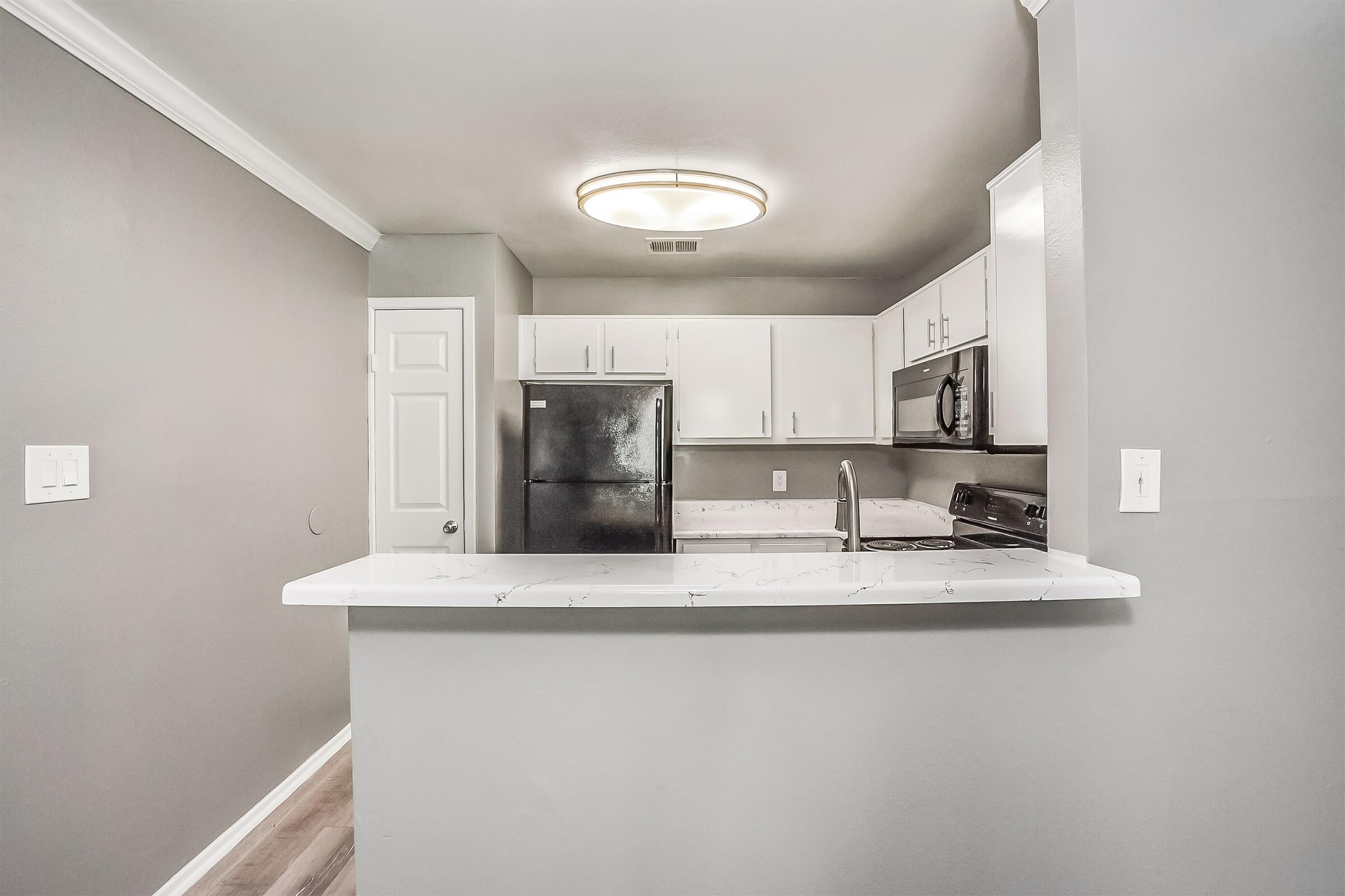 A modern kitchen featuring white cabinets, a black refrigerator, and stainless steel appliances. The kitchen island has a white countertop, and the walls are painted in a soft gray. A ceiling light fixture provides illumination, and the flooring is a light wood finish, creating a bright and inviting space.