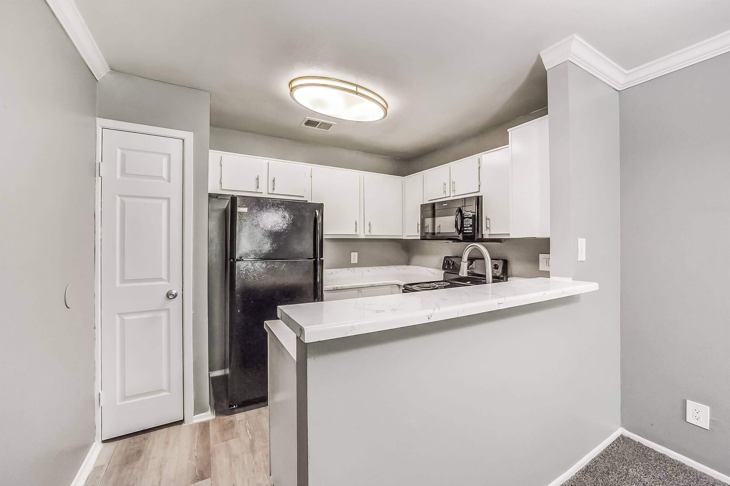 Modern kitchen featuring gray walls, a white countertop, and black appliances. The layout includes a refrigerator, stove, and overhead cabinetry. There's a small breakfast bar that separates the kitchen area from a living space, with natural light coming from an overhead fixture.