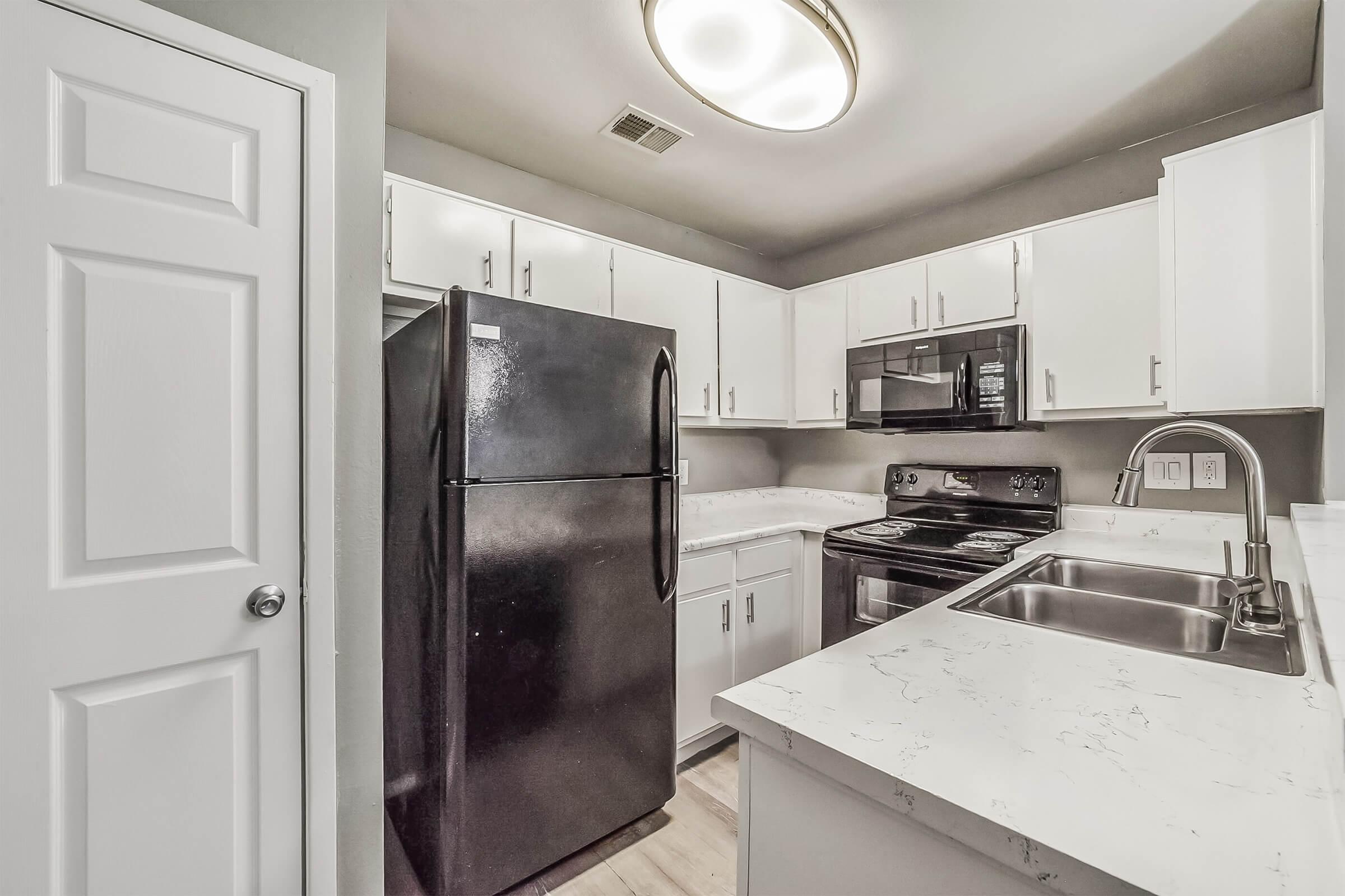 A modern kitchen featuring white cabinets, black appliances including a refrigerator and a stove, and a stainless steel sink. The countertops have a light, quartz-like appearance. A ceiling light fixture illuminates the space, and a door is visible on the left side. The overall design is clean and functional.
