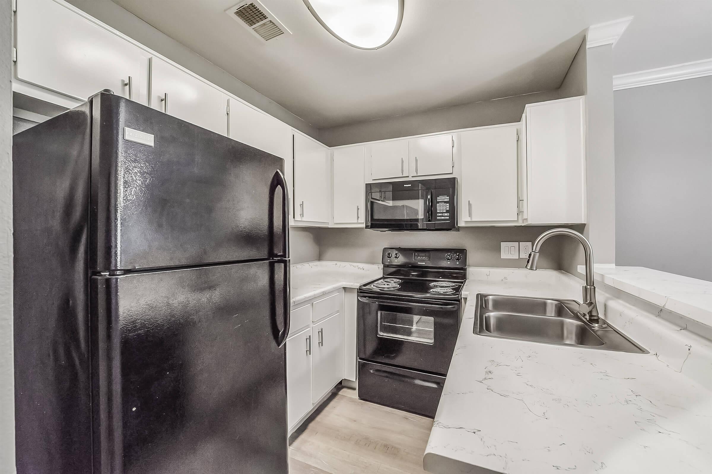 A modern kitchen featuring white cabinets, a black refrigerator, and an electric stove. The countertops are light-colored with a marbled design. There's a stainless steel sink and an overhead light fixture, creating a bright and functional cooking space. The walls are painted in a neutral gray tone.
