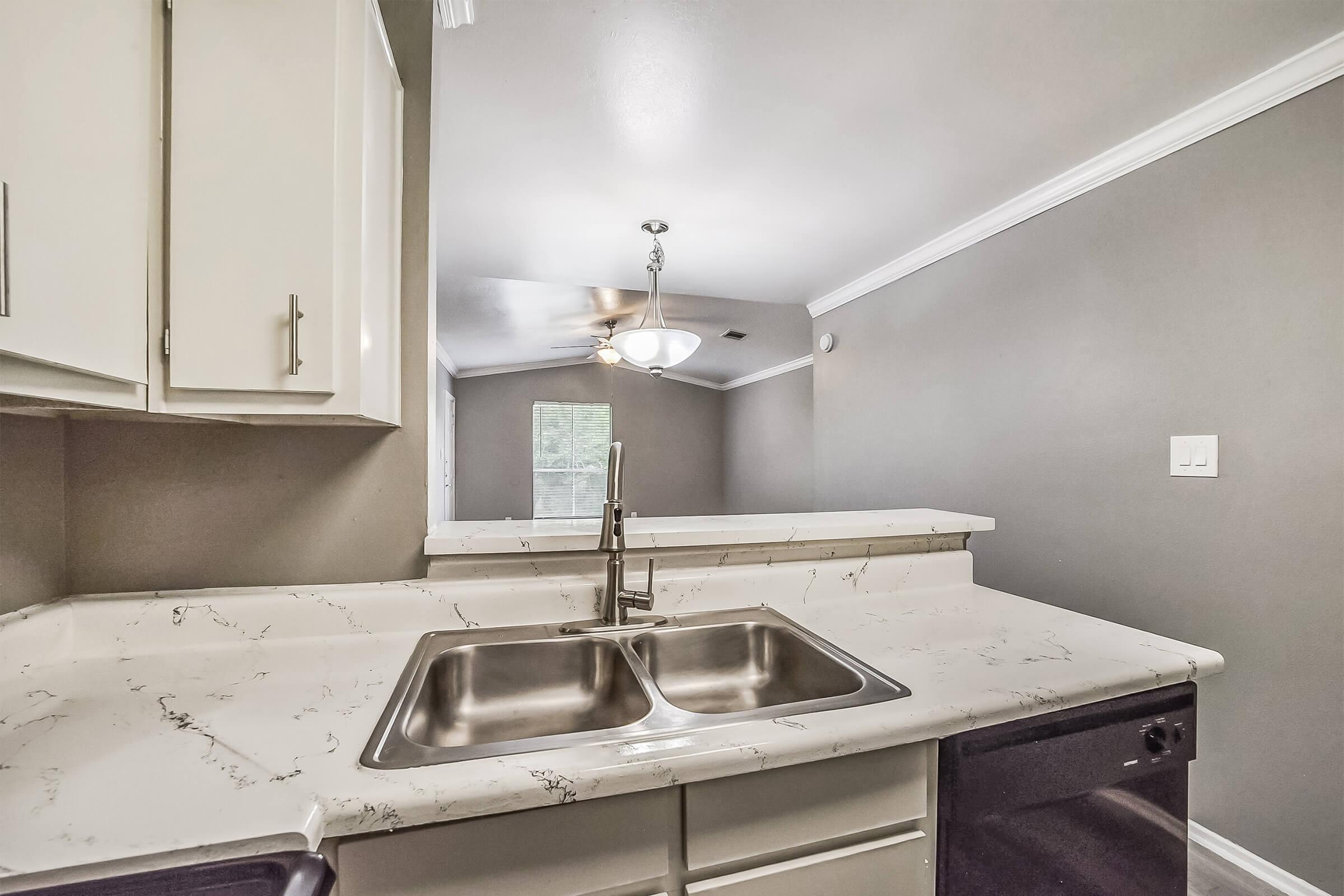 A modern kitchen featuring a double stainless steel sink, white cabinets with a marble-like countertop, and a black dishwasher. The background shows an open living space with a ceiling fan and neutral-colored walls, enhancing the airy feel of the area.