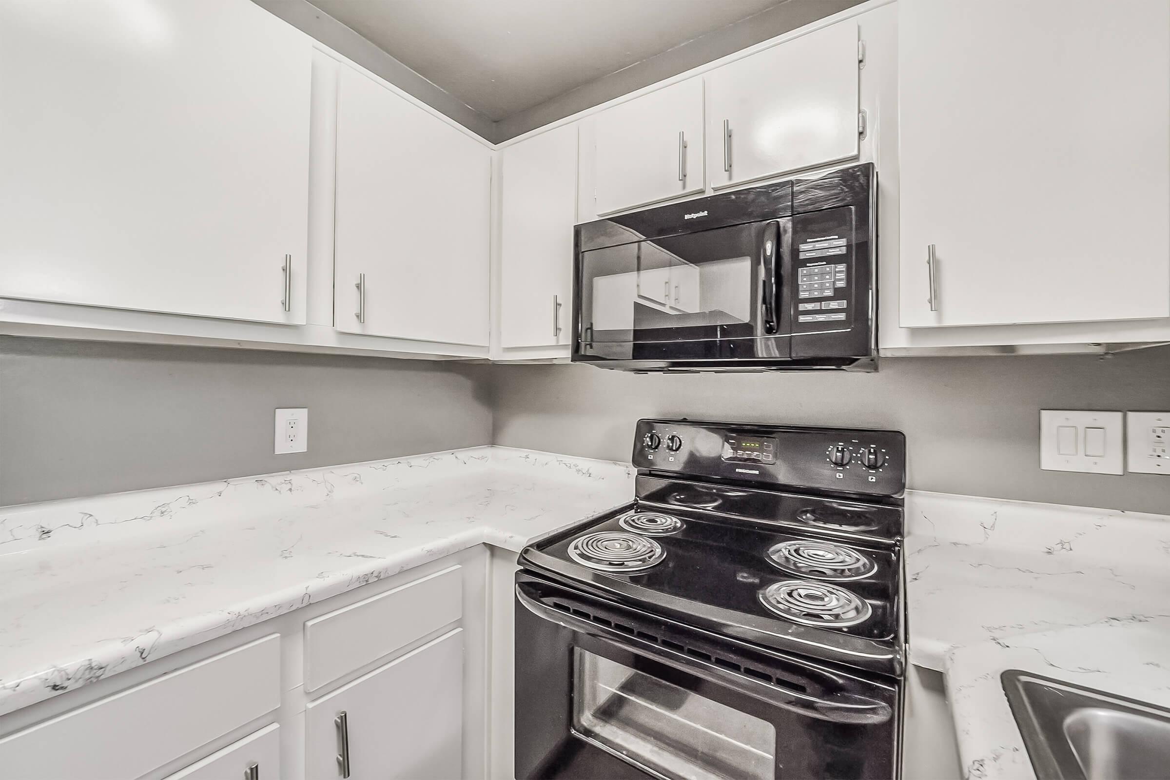 A modern kitchen featuring white cabinets, a marble-patterned countertop, a black stove, and a black microwave mounted above it. The walls are painted in a neutral gray, and there is a sink on the right side of the image. The overall design is clean and functional.