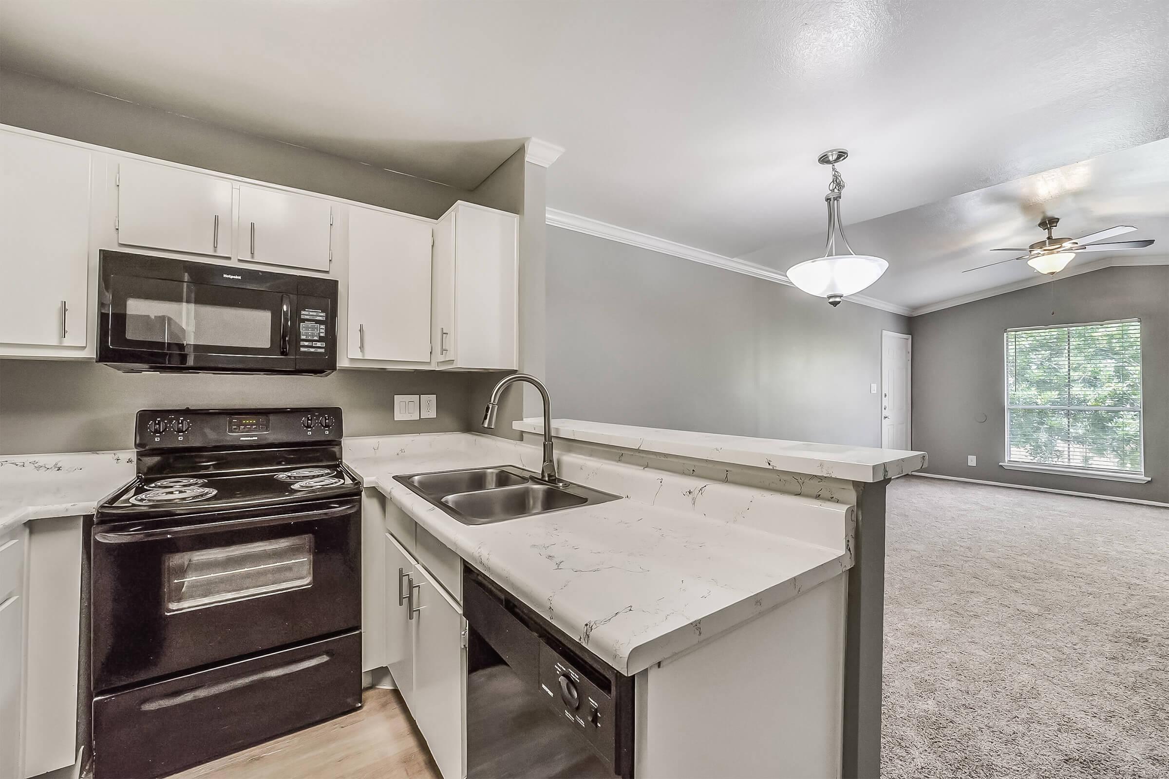 A modern kitchen featuring white cabinetry with a marble countertop, an integrated sink, and black appliances, including a stove and dishwasher. In the background, a spacious living area is visible, with carpeted flooring, a ceiling fan, and large windows allowing natural light.