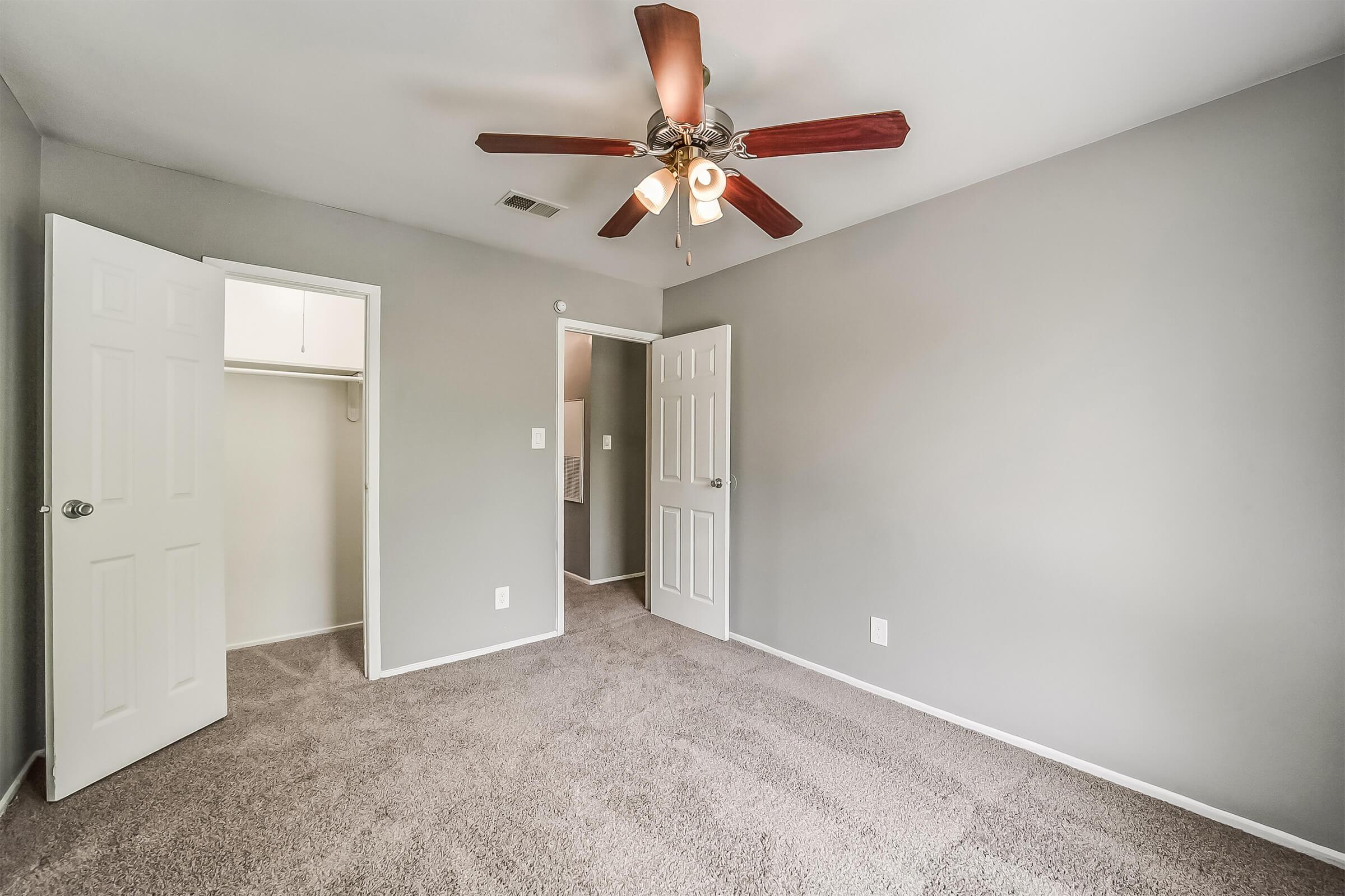 A well-lit, empty bedroom featuring gray walls and carpet. It has a ceiling fan with wooden blades, a closet door on the left, and an open white door leading to another room. The overall atmosphere is clean and modern, creating a spacious feel.