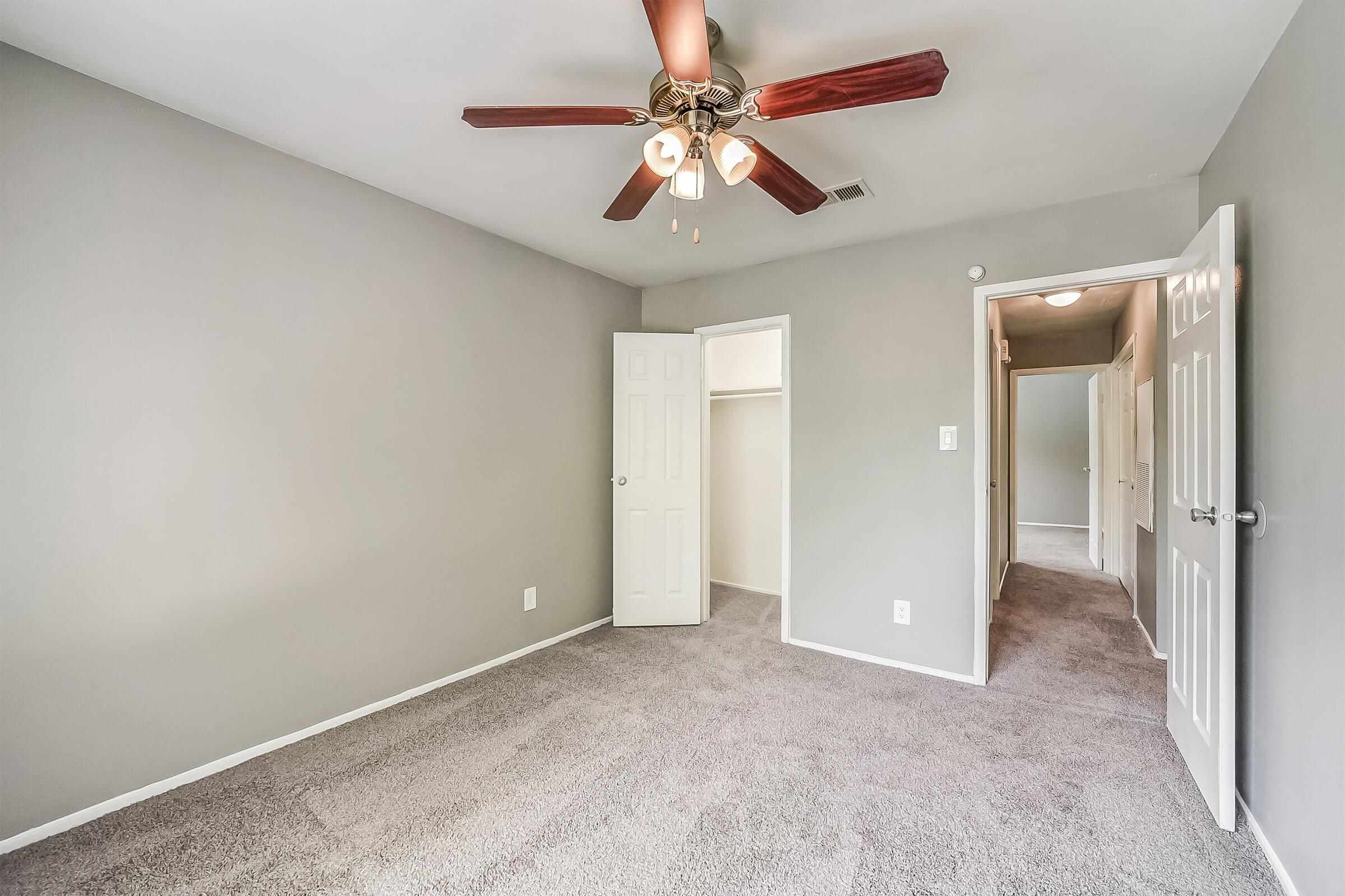 A neutral-colored bedroom featuring gray walls and carpeting, a ceiling fan with wooden blades, and two doors—one leading to a closet and another to a hallway. The room is well-lit and minimalistic, with a clean and spacious feel.
