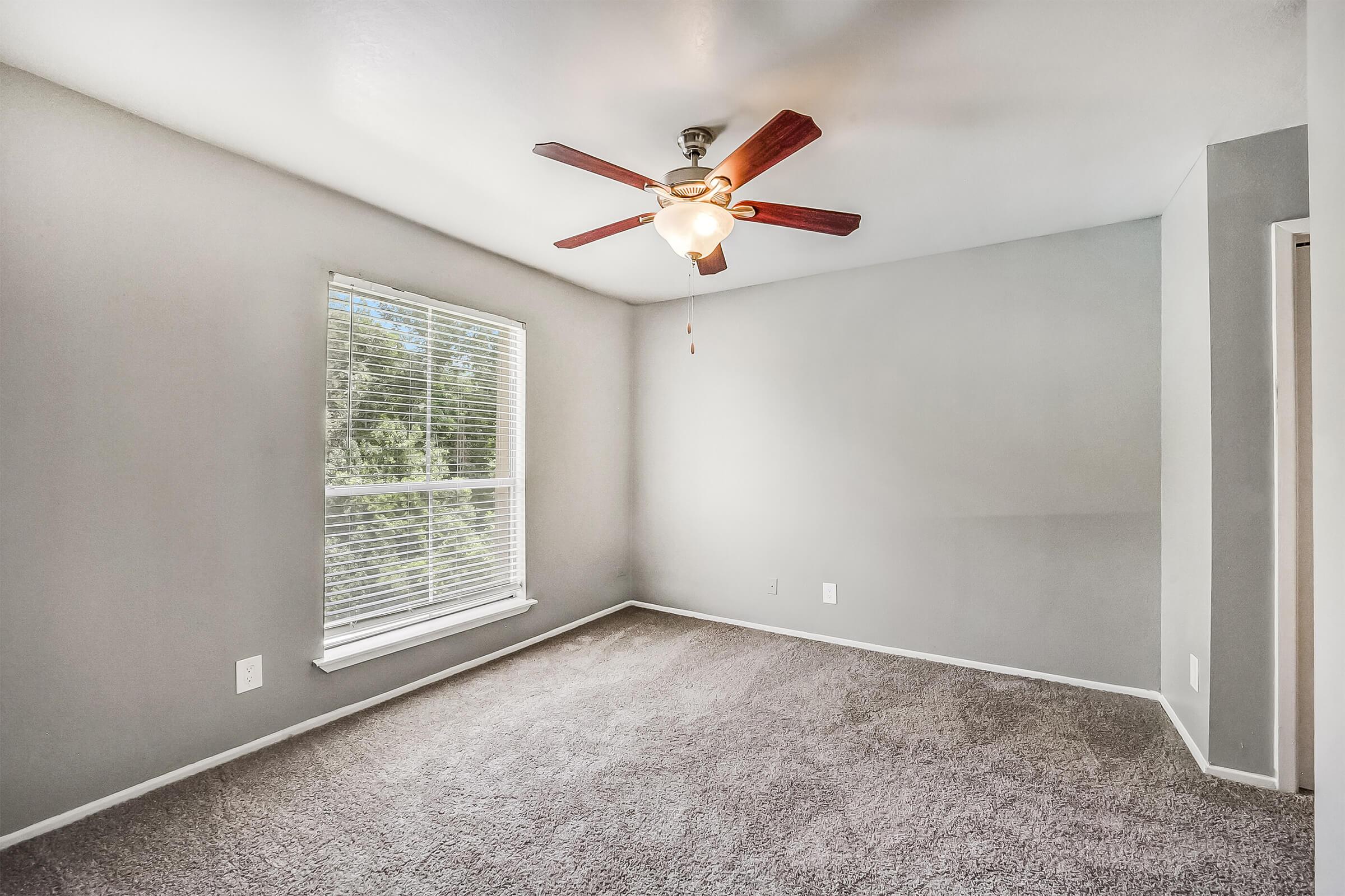 A spacious, empty room with light gray walls and a ceiling fan with wooden blades. There is a large window on one wall, allowing natural light to illuminate the soft, carpeted floor. The room has a minimalist design, with no furniture or decorations, creating an inviting atmosphere.