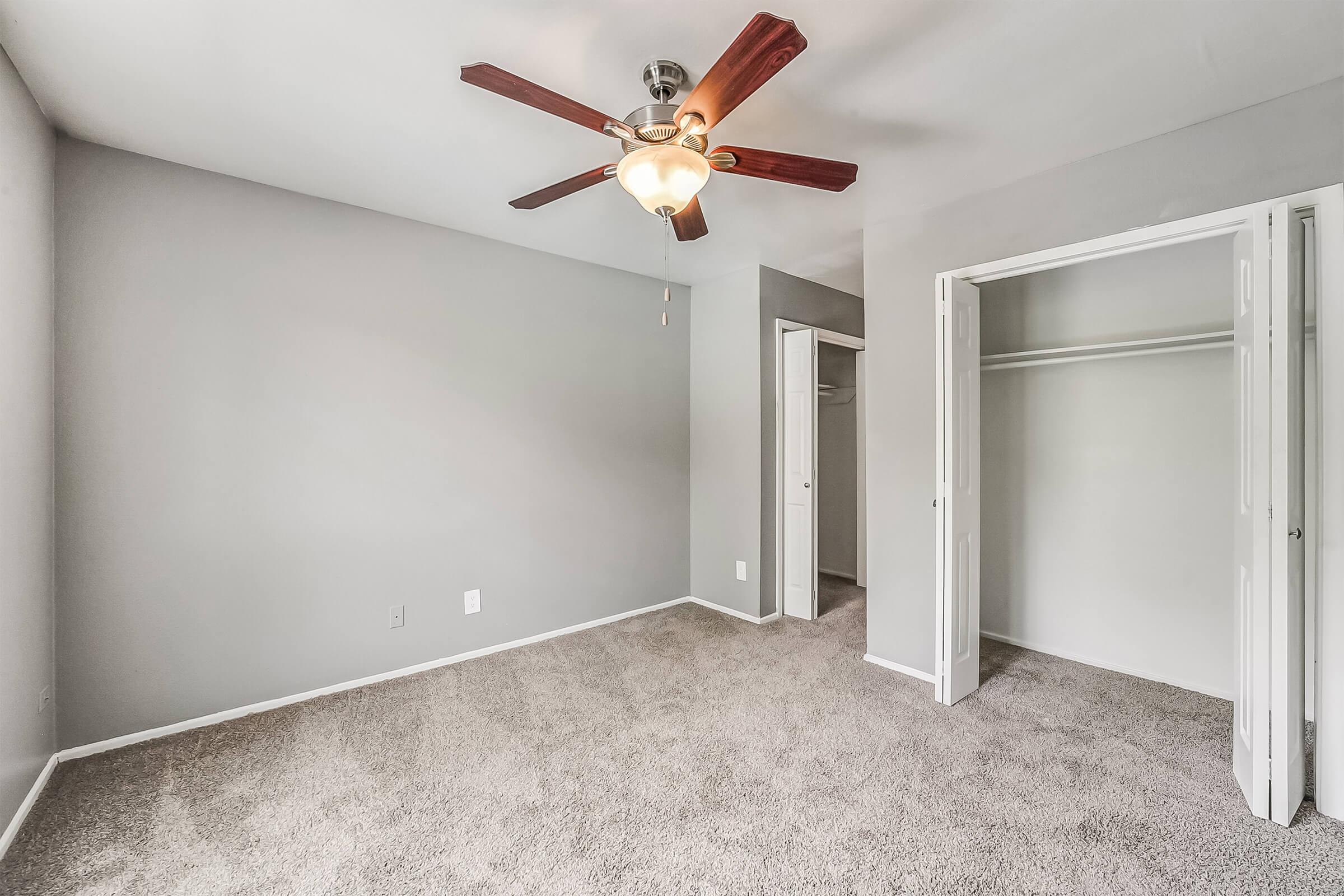 A spacious, unoccupied bedroom featuring light gray walls and plush carpet. A ceiling fan with wooden blades hangs in the center, while a closet with sliding doors is visible to the right. The room is well-lit with natural light, creating an airy atmosphere.