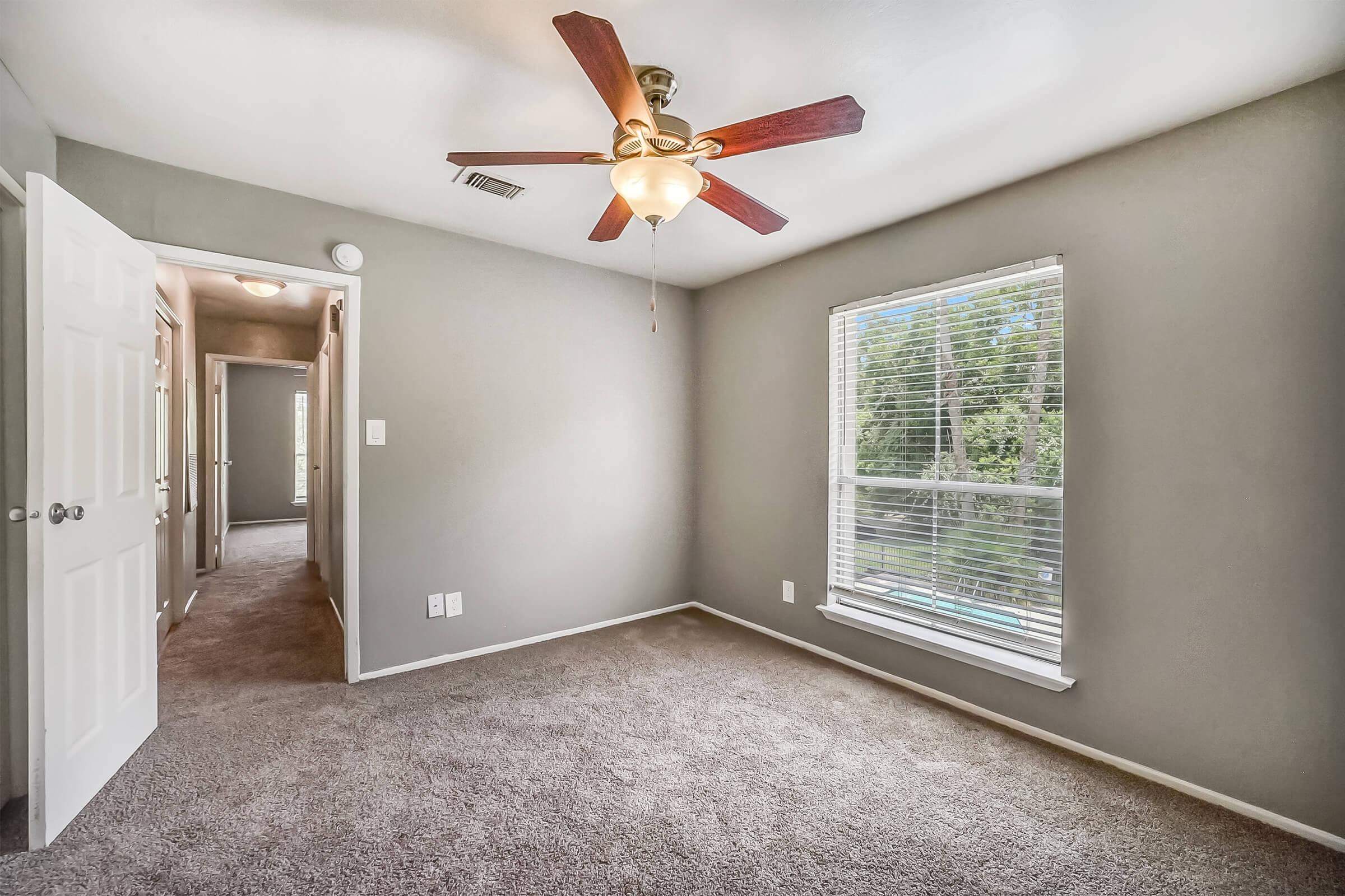 A well-lit room with a ceiling fan and light fixture, featuring gray walls and carpeted flooring. A window with blinds allows natural light, showing greenery outside. An open door leads to a hallway, indicating additional space beyond the room.