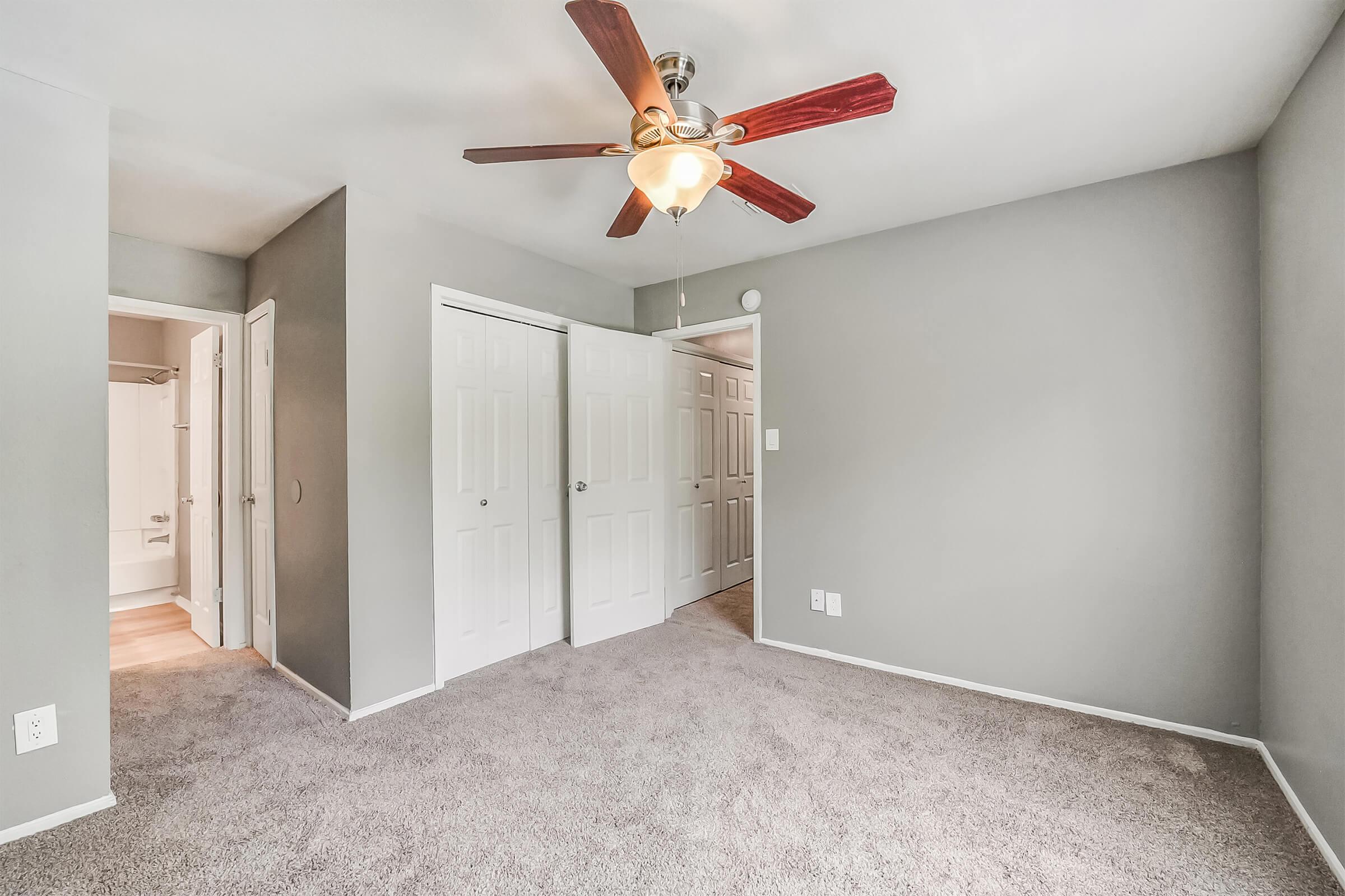 A spacious, light-filled bedroom featuring gray walls and plush carpet. The room includes a ceiling fan with wooden blades, a large window, and two closed doors leading to a closet and a bathroom. The overall atmosphere is modern and inviting.