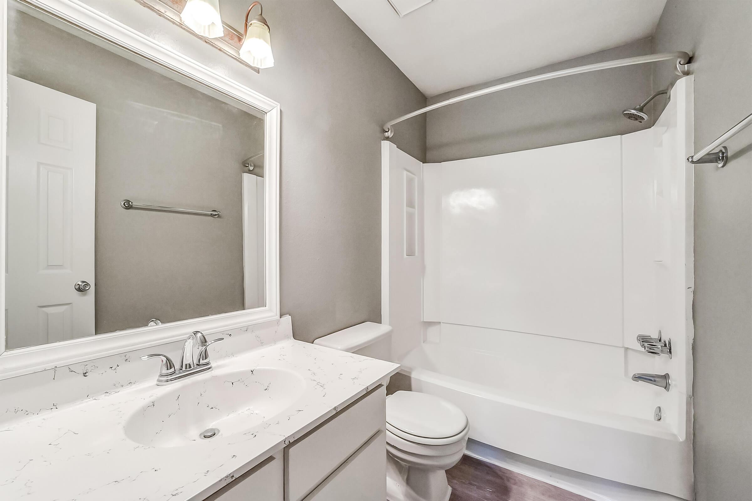 A clean and modern bathroom featuring a white bathtub with a curved shower curtain rod, a sleek white sink with a marble countertop, a large mirror above the sink, and gray walls. There is a door on the left leading to another space and a towel rack on the wall.