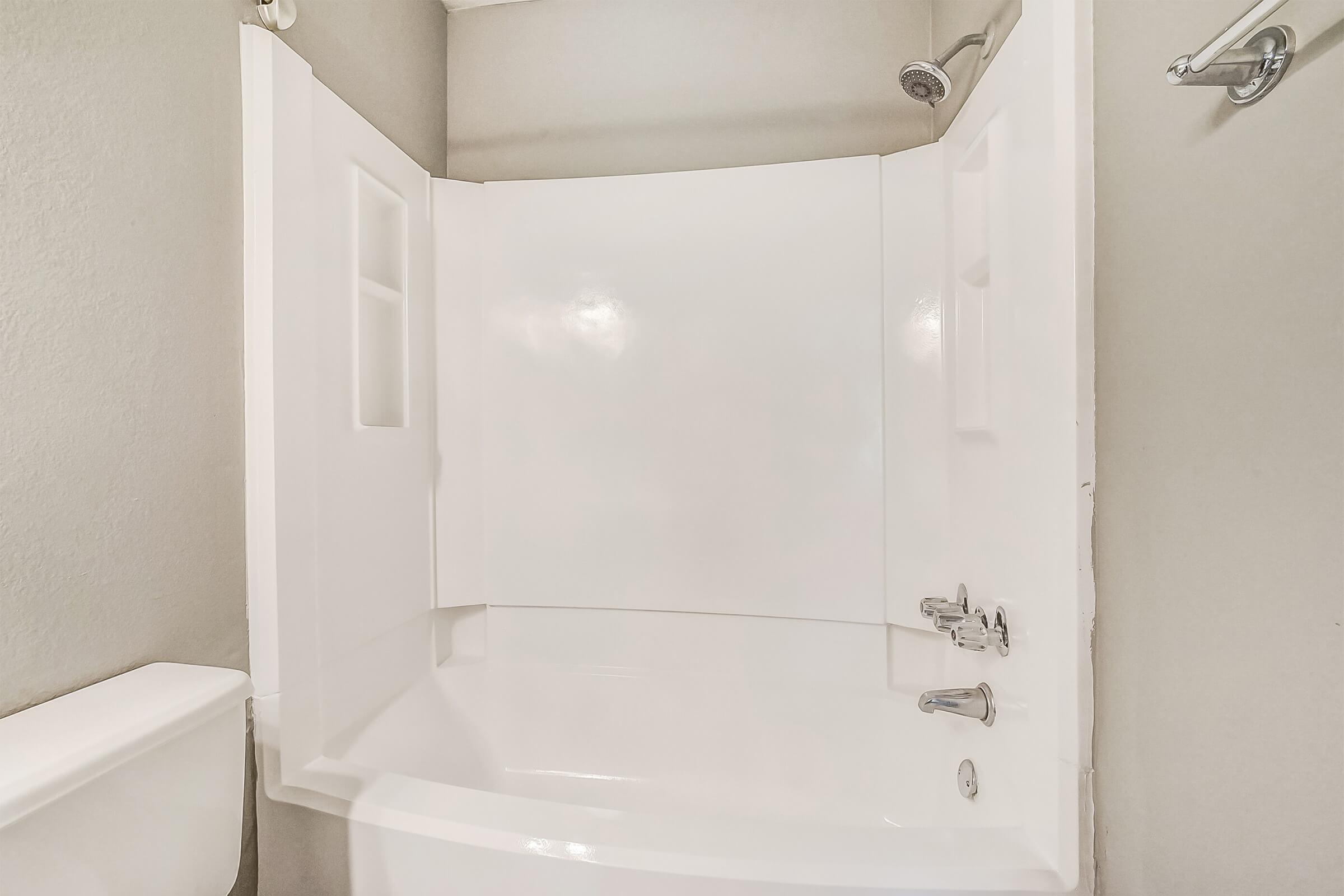 A modern bathroom featuring a clean, white tub and shower combination with smooth walls. The tub has a chrome faucet and showerhead, and there are built-in shelves on the wall for toiletries. The background is a neutral gray, contributing to a minimalist aesthetic.