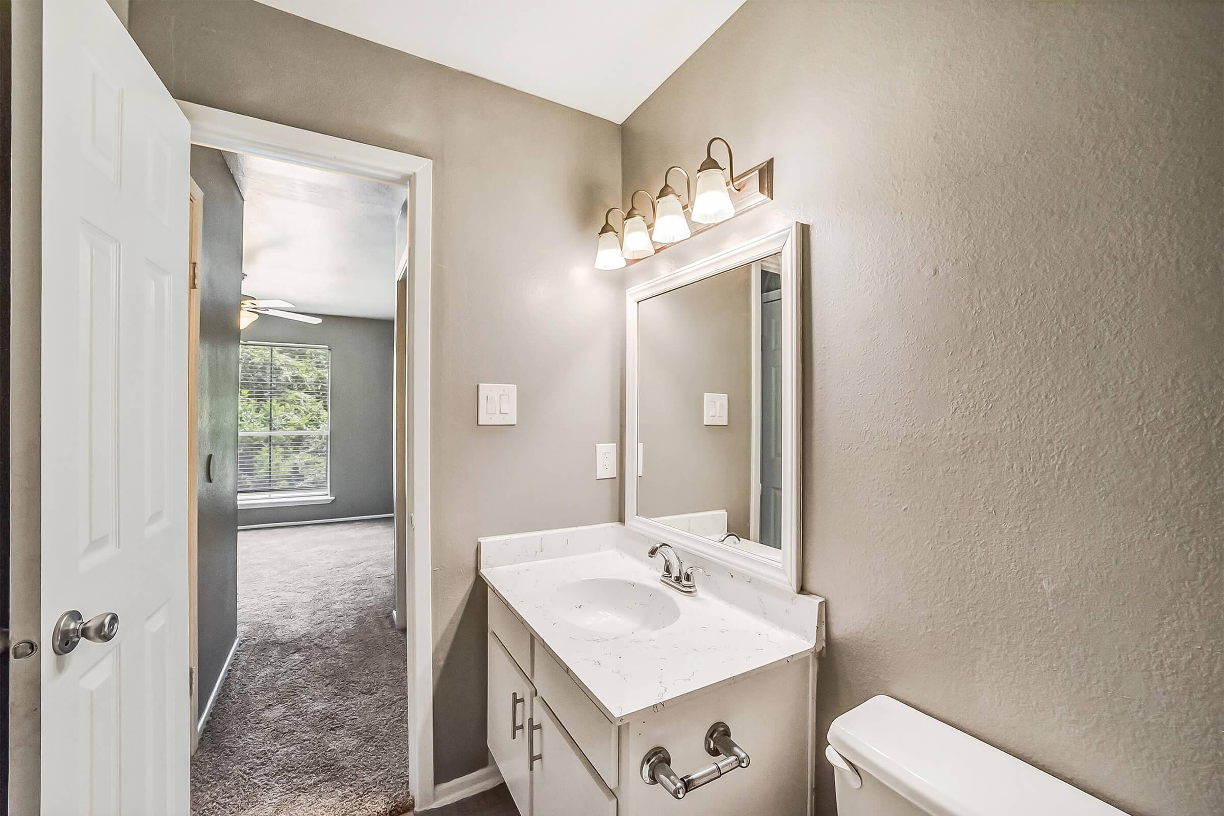 A bright bathroom featuring a light gray color scheme. It includes a white countertop with a sink, a large mirror above, and a modern light fixture. A door leads to another room, while a window offers natural light. The floor is carpeted, contrasting the clean surfaces.