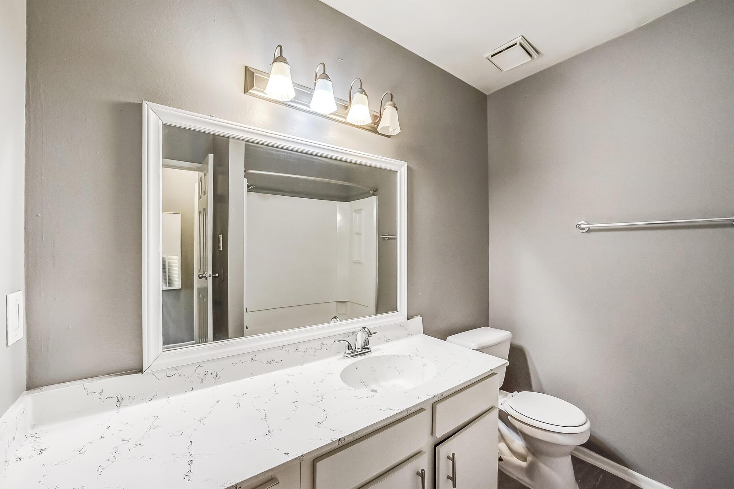 A modern bathroom featuring a white marble countertop with a sink, a large mirror, a multi-light fixture above, a toilet, and a shower area. The walls are painted in a soft gray color, creating a clean and contemporary look.
