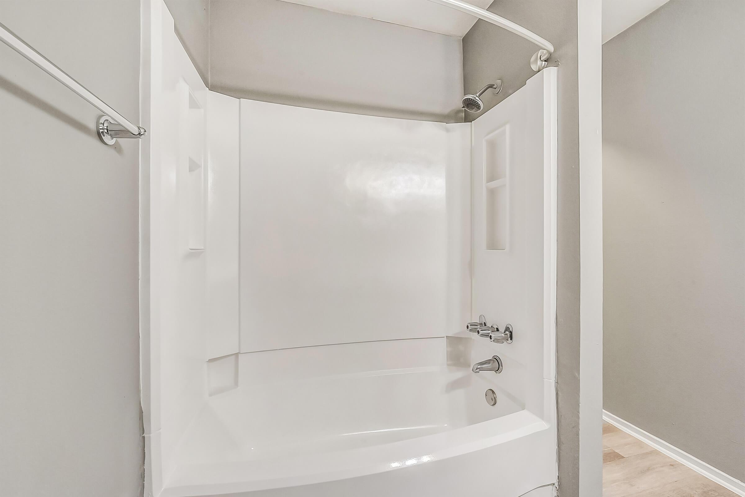 A clean, modern bathroom featuring a white fiberglass shower tub combination. The walls are painted in a light gray color, and there are two built-in shelves for storage. A chrome showerhead and faucet are visible, along with a curved shower rod. The flooring appears to be a light wood or laminate.