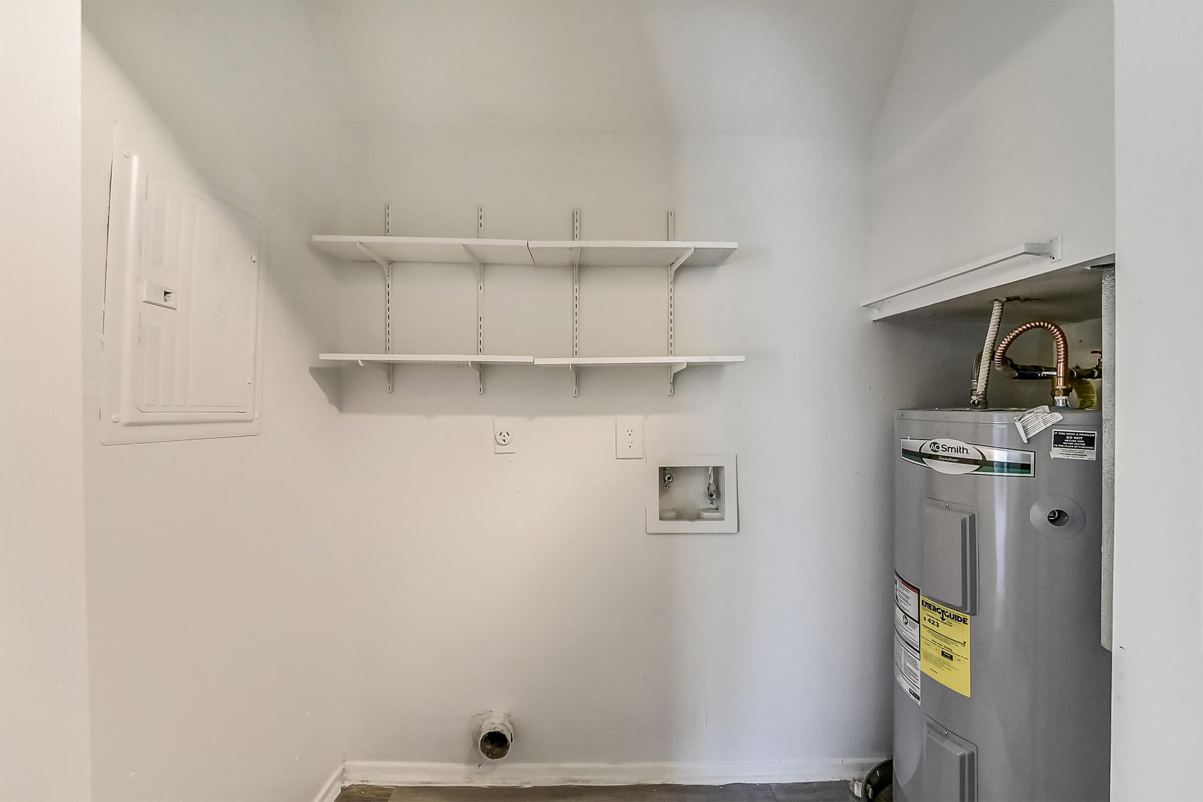 Small laundry room with white walls featuring a water heater, a pipe, and wall-mounted shelves. The floor is bare, and there is a small access panel on the left side. The overall space appears clean and functional, suitable for laundry appliances or storage.