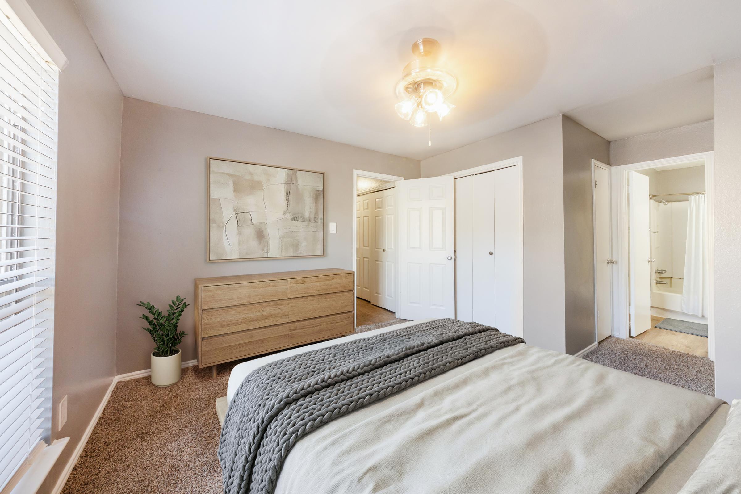 A cozy bedroom featuring a bed with a textured blanket, a wooden dresser, and a potted plant. The walls are painted in a light shade, and there are two doorways leading to other rooms. Natural light filters through the window with white blinds, creating a warm and inviting atmosphere.