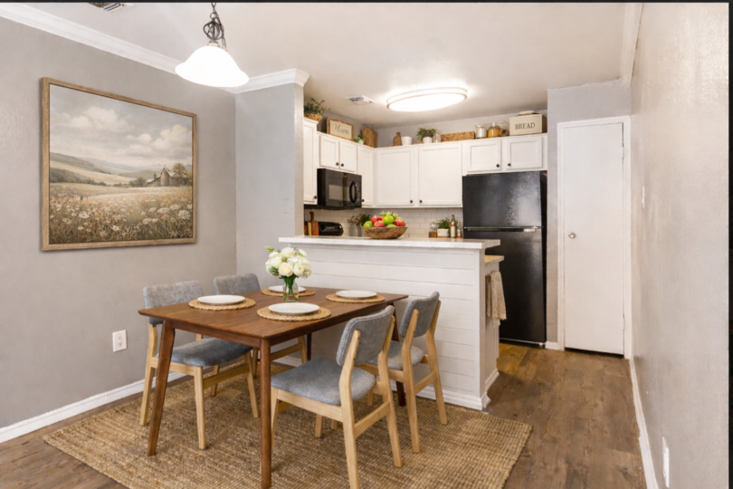 A cozy dining area featuring a wooden dining table set for four, surrounded by light gray chairs. The room has a light gray wall with a landscape painting, a modern light fixture, and a kitchen with white cabinetry and a black refrigerator. A woven rug adds texture to the space.