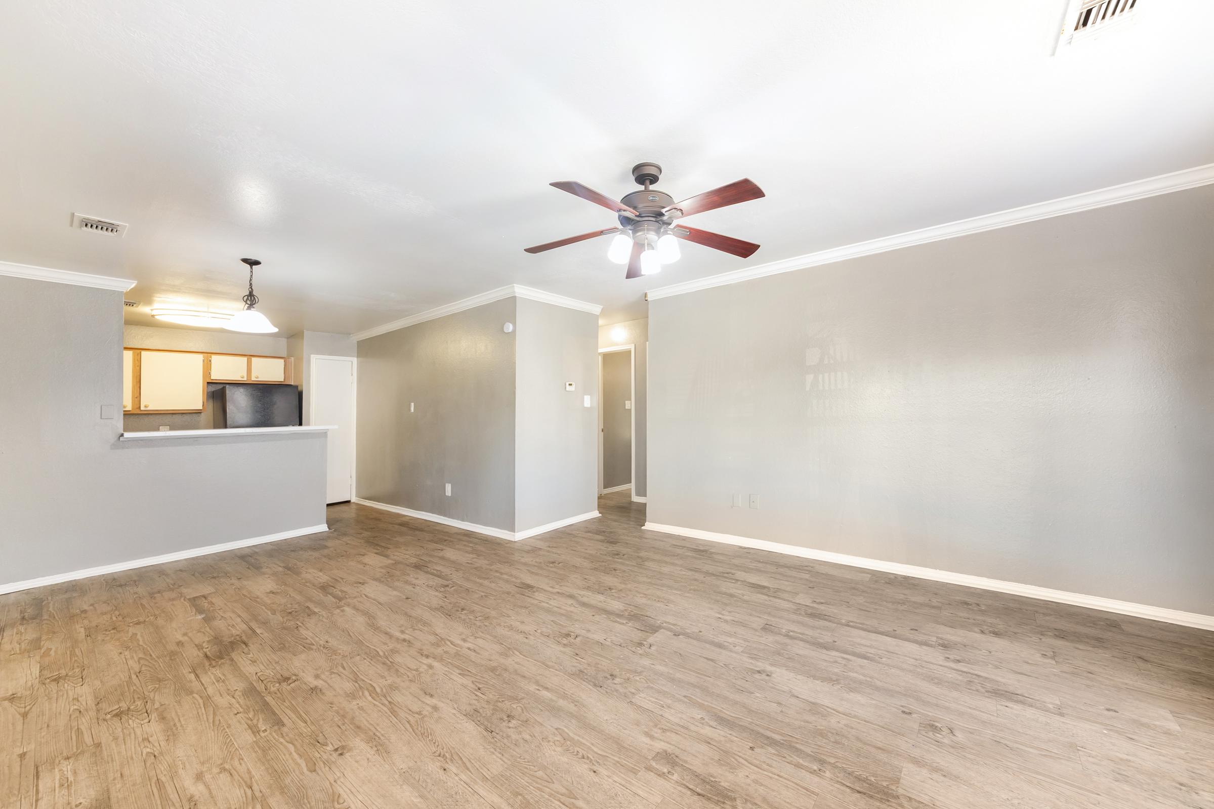 A spacious living area featuring light-colored walls and a ceiling fan. The floor is covered in wood-like laminate. In the background, there's a kitchen with an island counter and a doorway leading to another room. Natural light enhances the open and airy feel of the space.