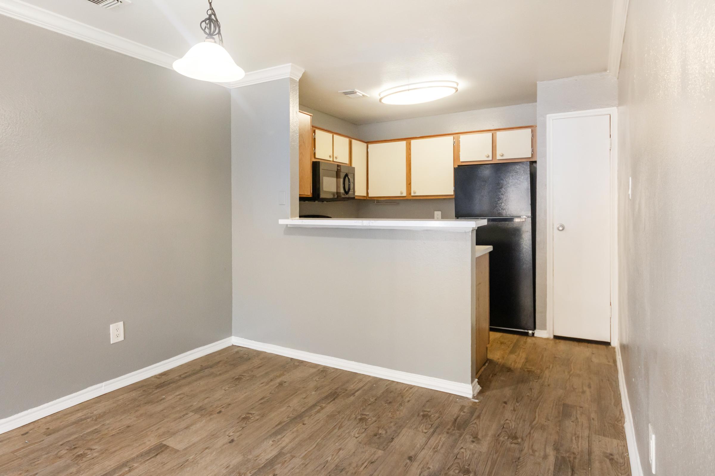 A modern kitchenette with wood-style flooring, featuring a bar counter, black refrigerator, and wooden cabinets. The walls are painted light gray, and there is a circular ceiling light. A door leads to another room, and the space is well-lit and uncluttered, providing a minimalist aesthetic.