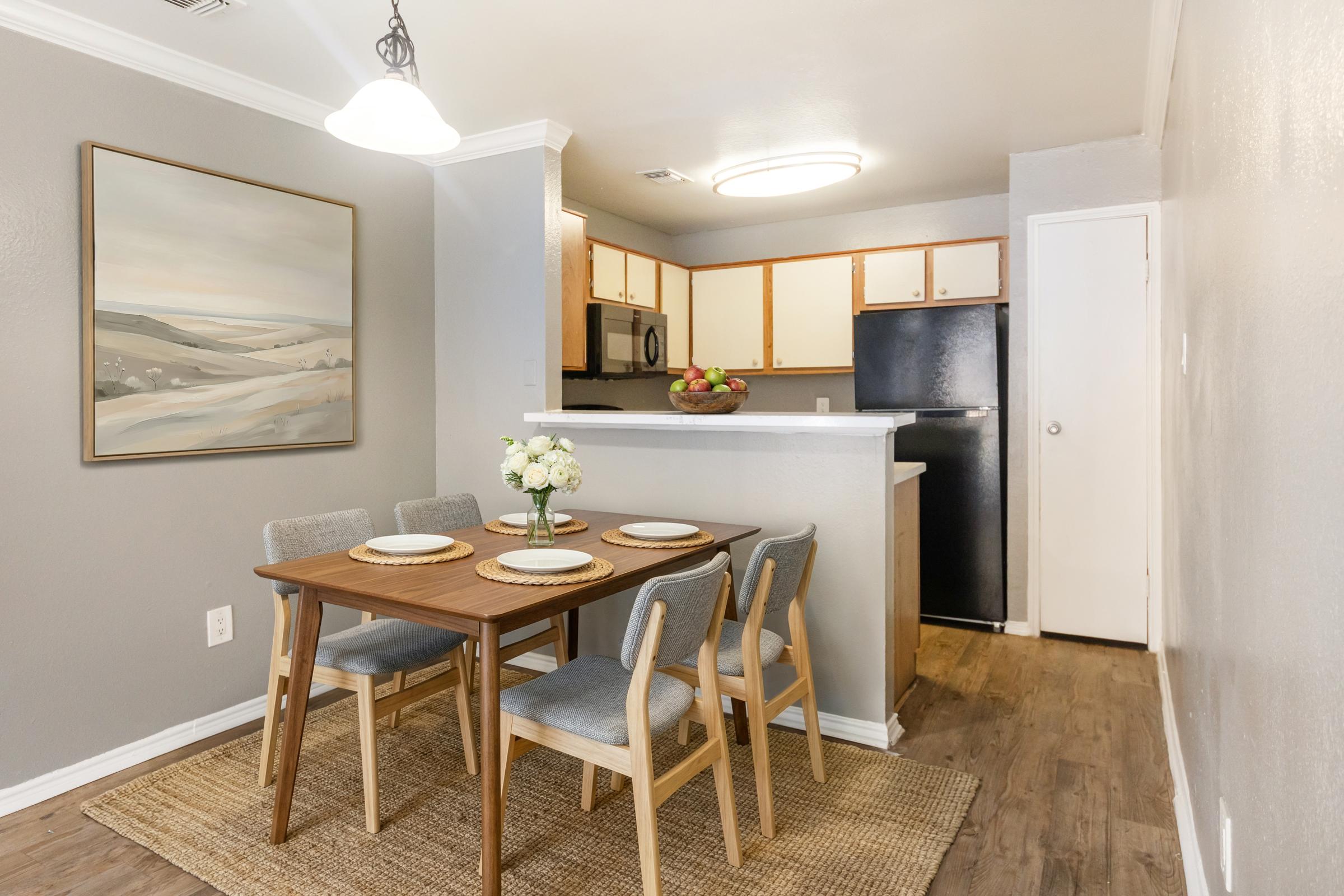 A cozy dining area featuring a wooden table set for four with floral centerpiece, surrounded by modern chairs. The space is decorated with a landscape painting, and includes an open kitchen with wooden cabinets, a black refrigerator, and stainless steel appliances. The flooring is light-colored wood, enhancing the inviting atmosphere.