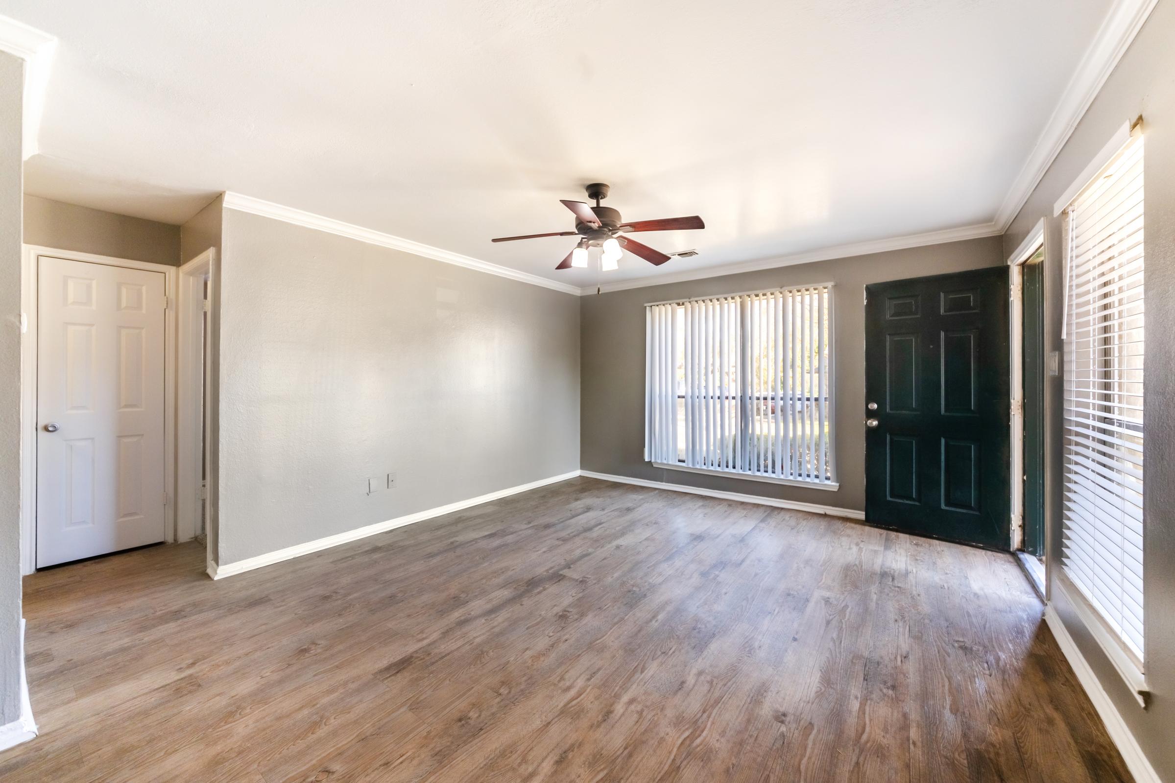 A spacious living room featuring light-colored walls, a ceiling fan, and large windows with vertical blinds. The floor has a wood-like finish, and there is a dark-colored door leading outside. A hallway with a closed door is visible on the left side.